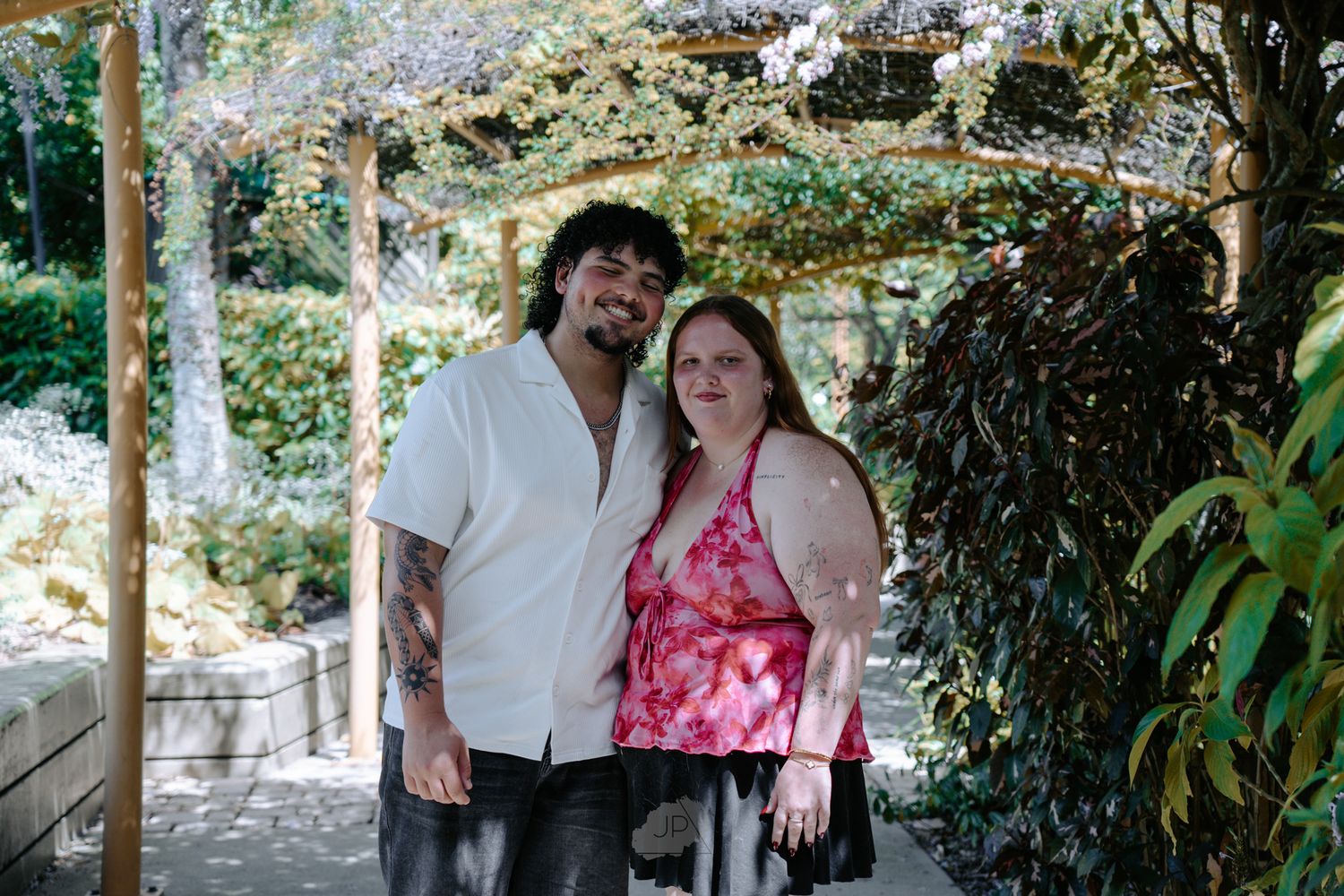 A couple shares a romantic moment in a lush garden setting with trees and greenery in the background.