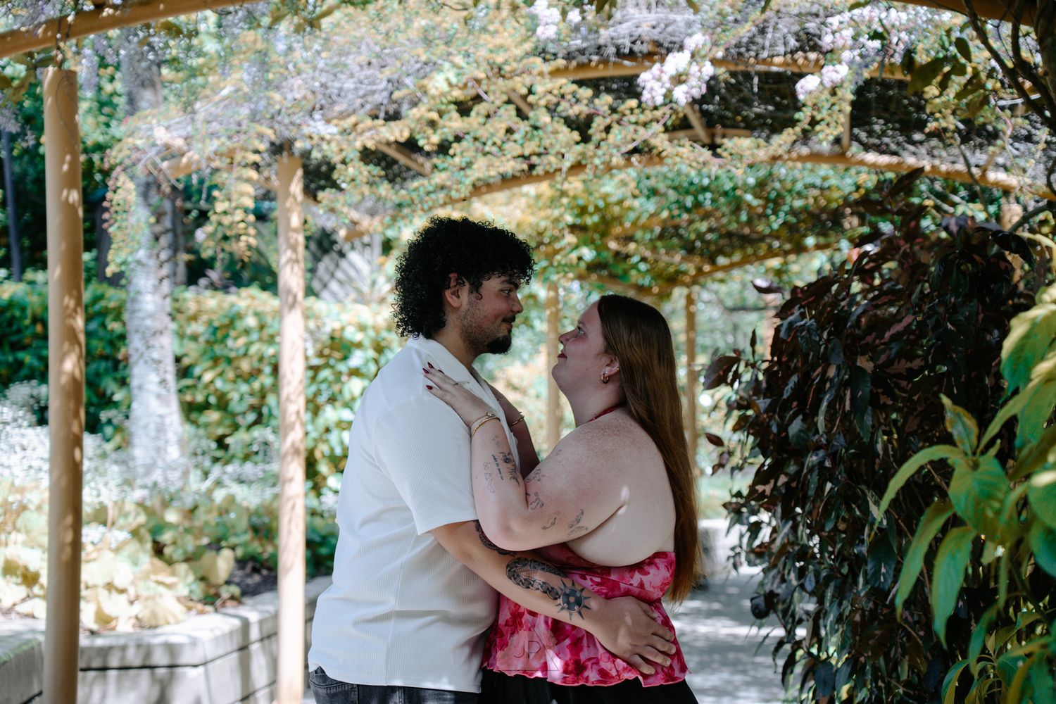 A couple shares a romantic moment under a white gazebo surrounded by lush greenery and flowers in a garden setting.
