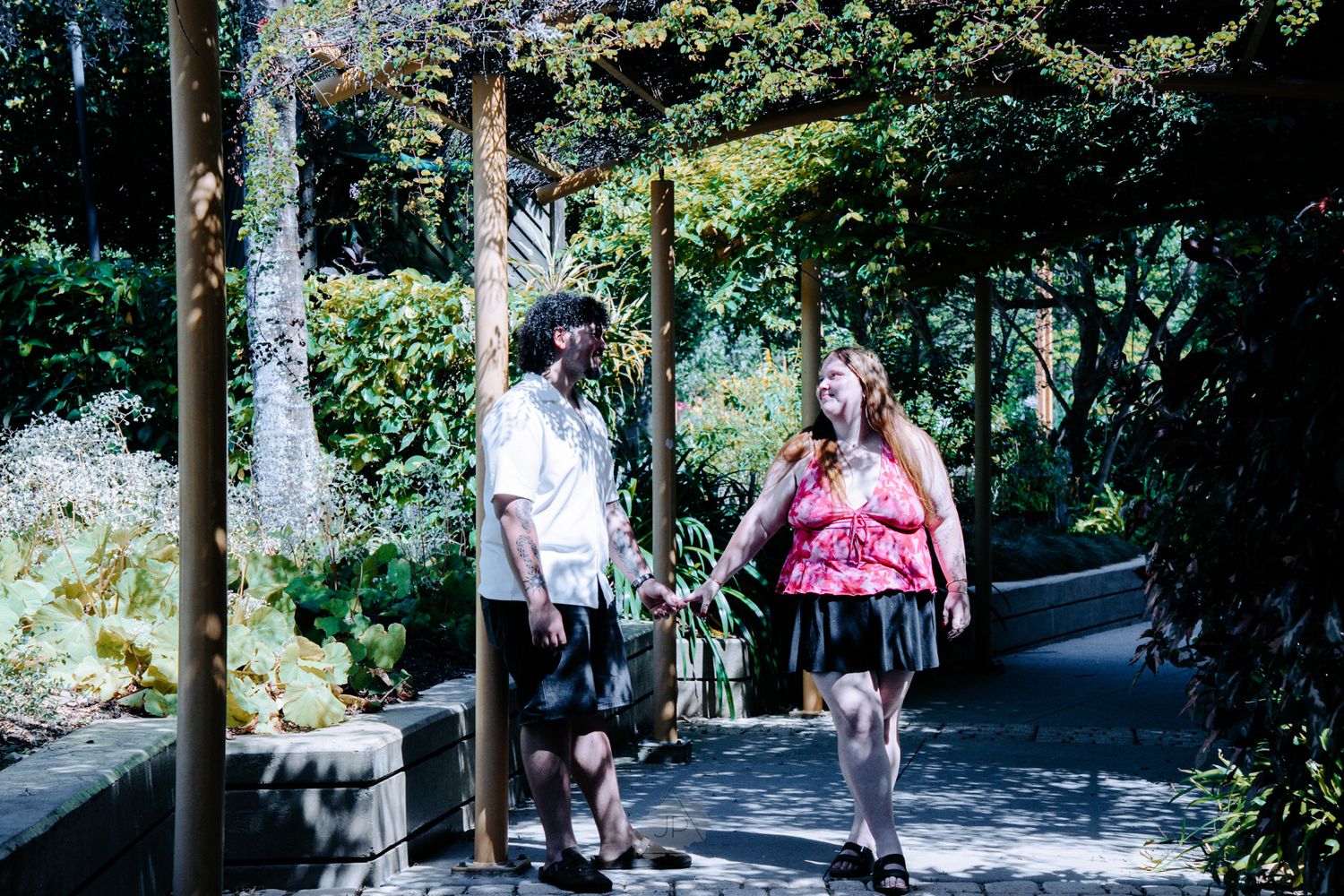 Two people walking dogs on a paved path through a lush green garden on a sunny day.