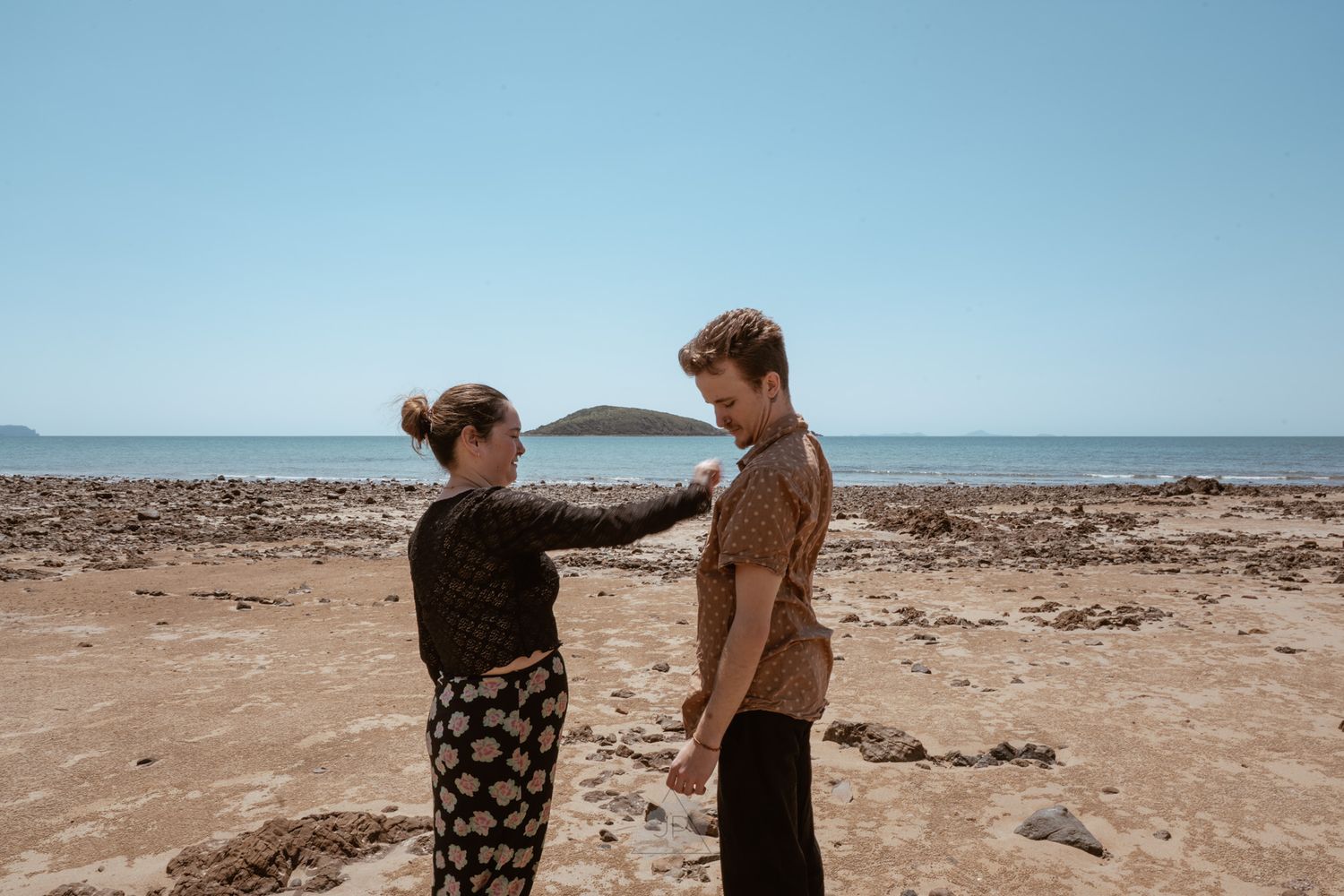Two people explore a barren coastal landscape with an island visible in the background.