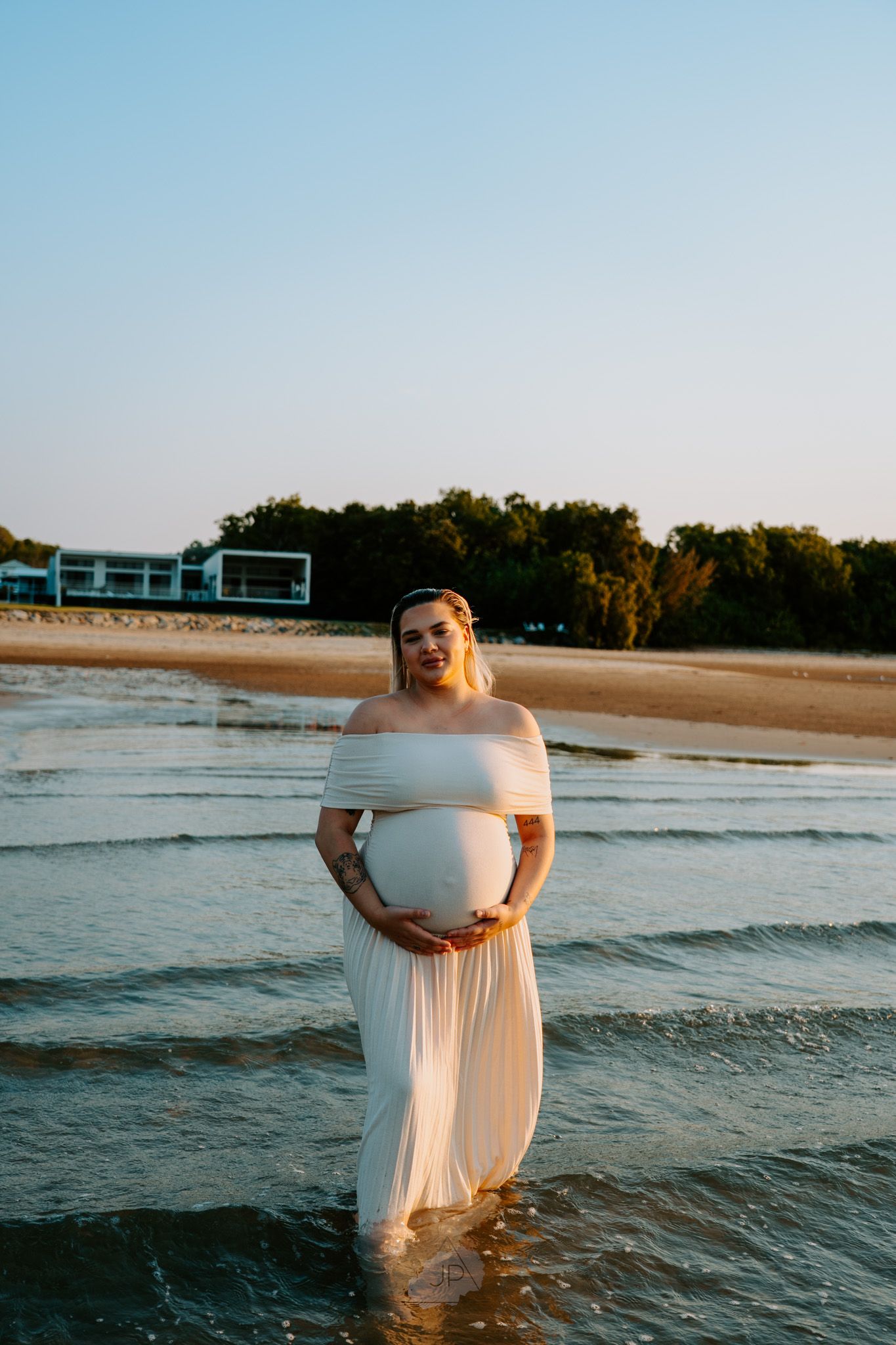 A figure in a flowing white dress stands in ocean waves at sunset creating a dreamy beachside scene.