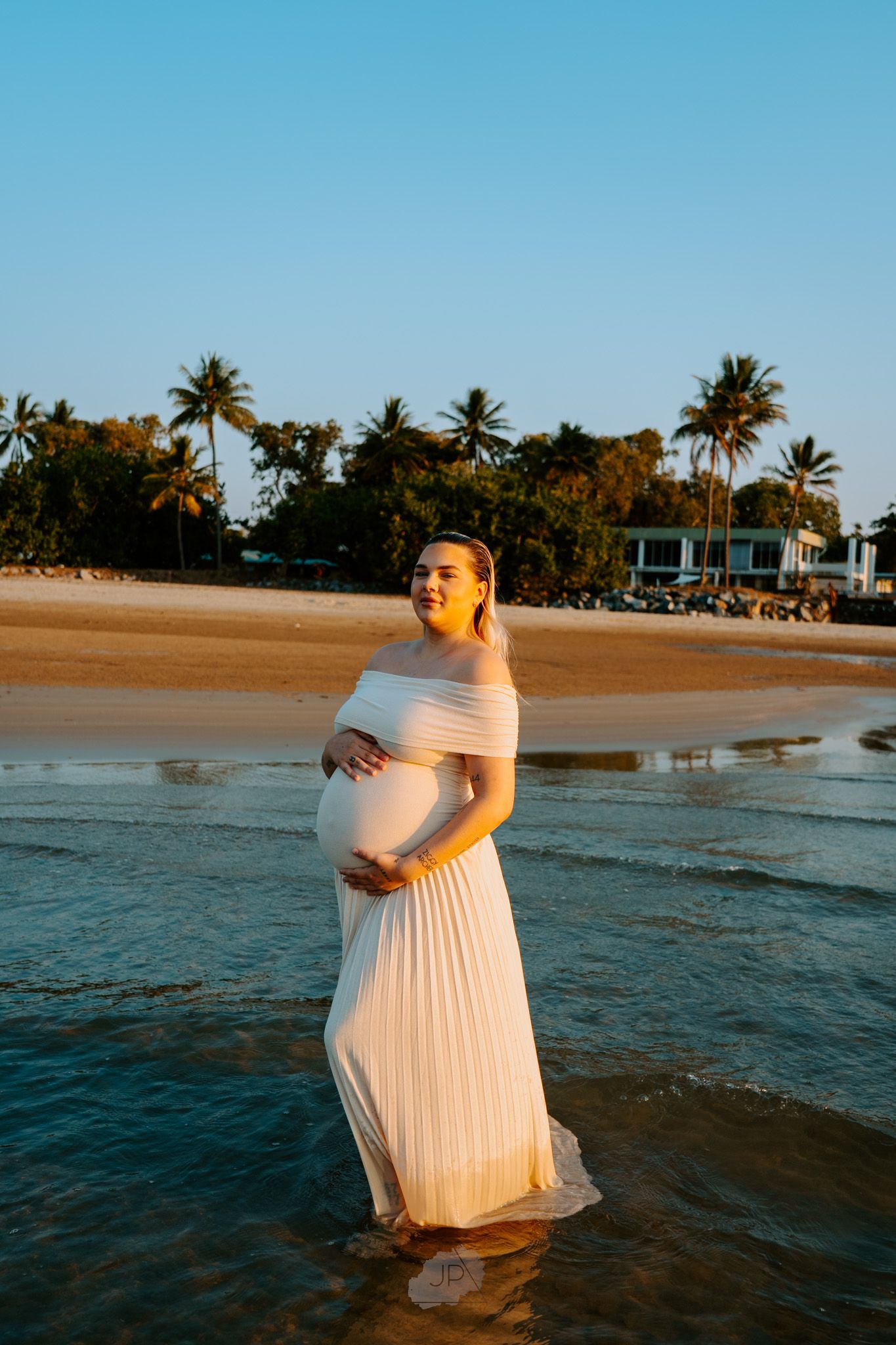 A person wearing a white dress poses in the ocean waves at golden hour with a tropical beach backdrop.
