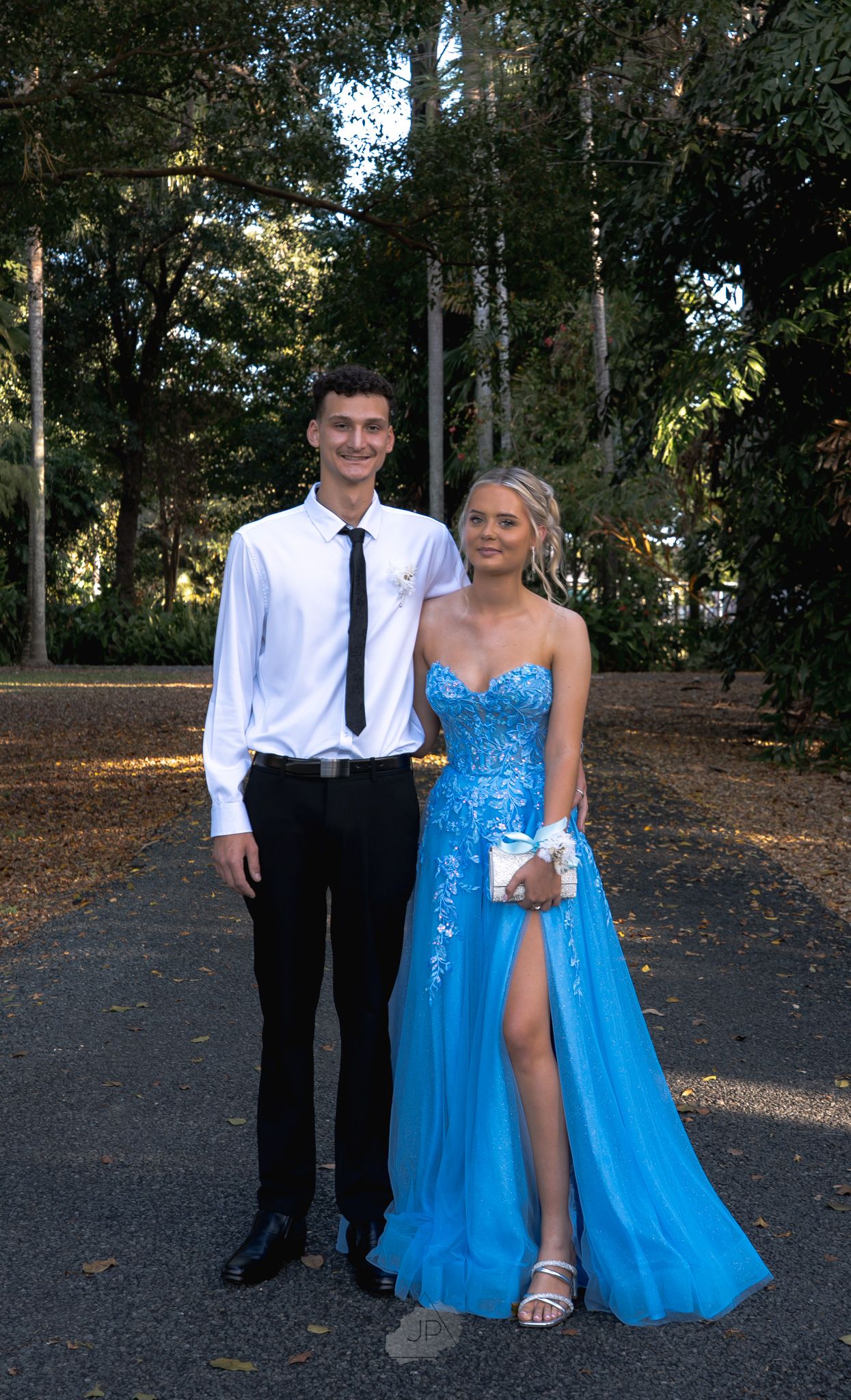 A couple poses together outdoors in formal attire with the person in a bright blue sparkly gown and white dress shirt and tie.