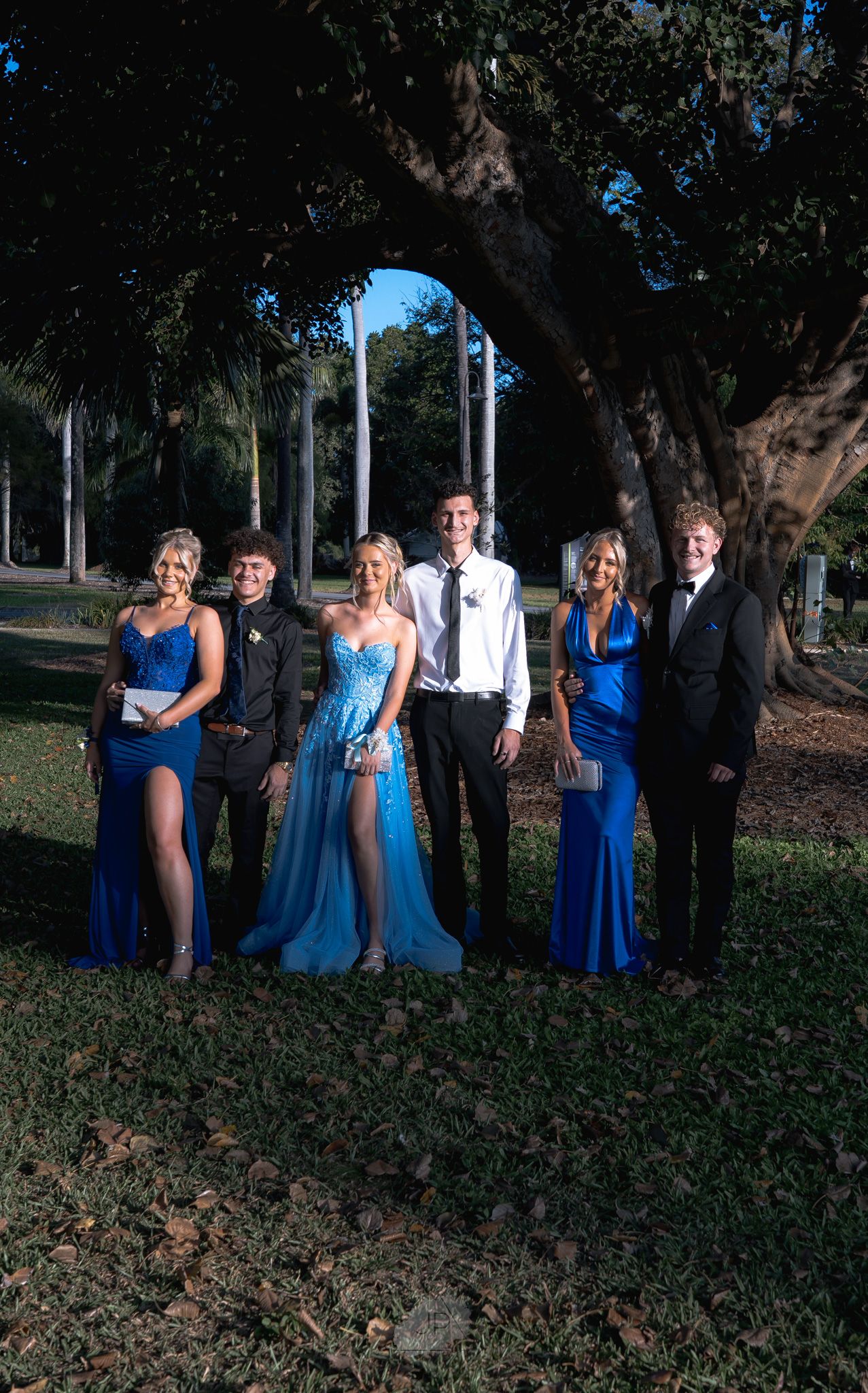 Wedding party members in coordinated blue dresses and suits stand together in a shaded outdoor location.
