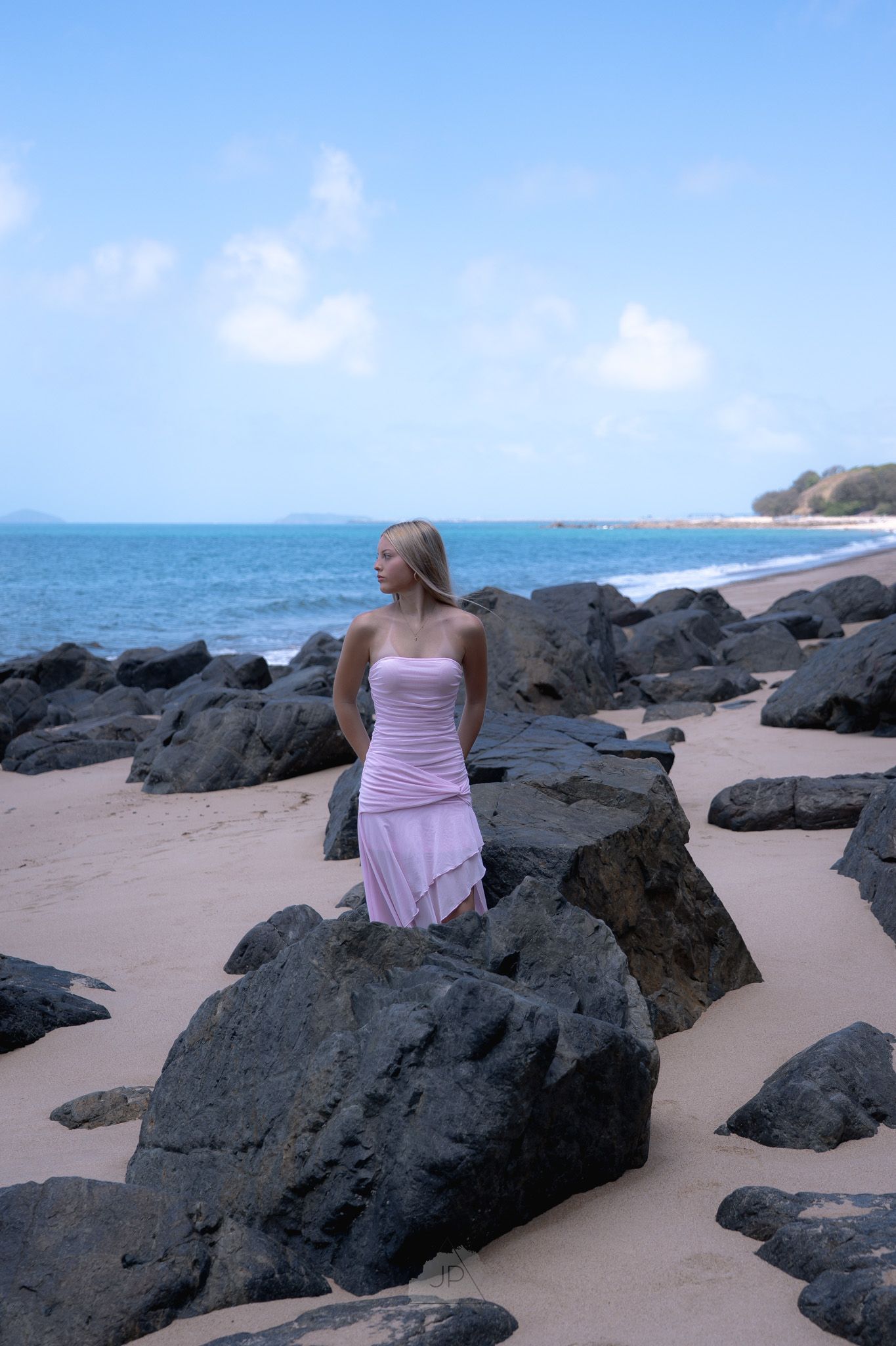 A person in a pink dress sits on dark volcanic rocks along a sandy beach with blue ocean waters in the background.