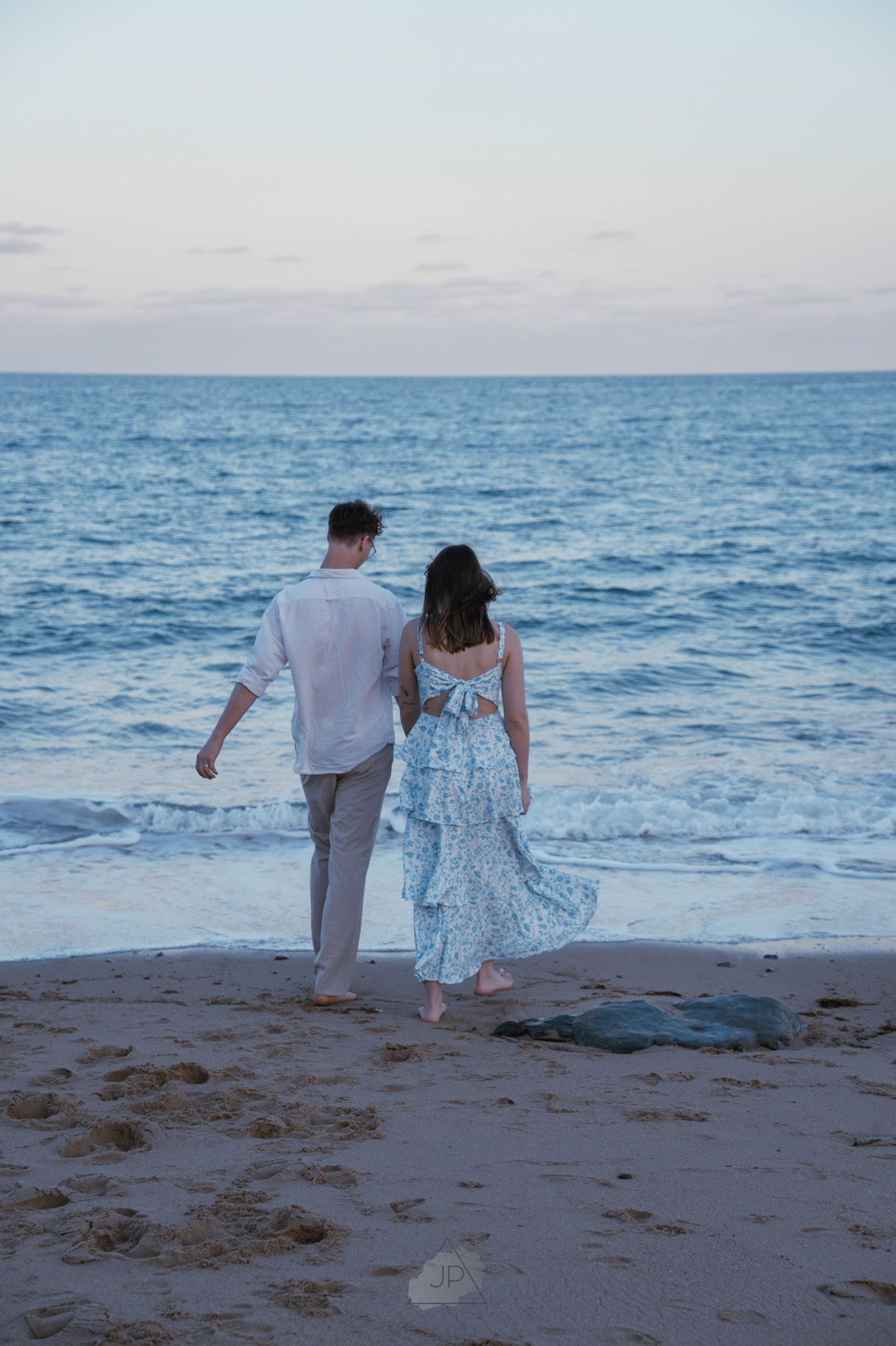 A couple in light colored outfits walks hand in hand along a serene beach at dusk with gentle waves in the background.