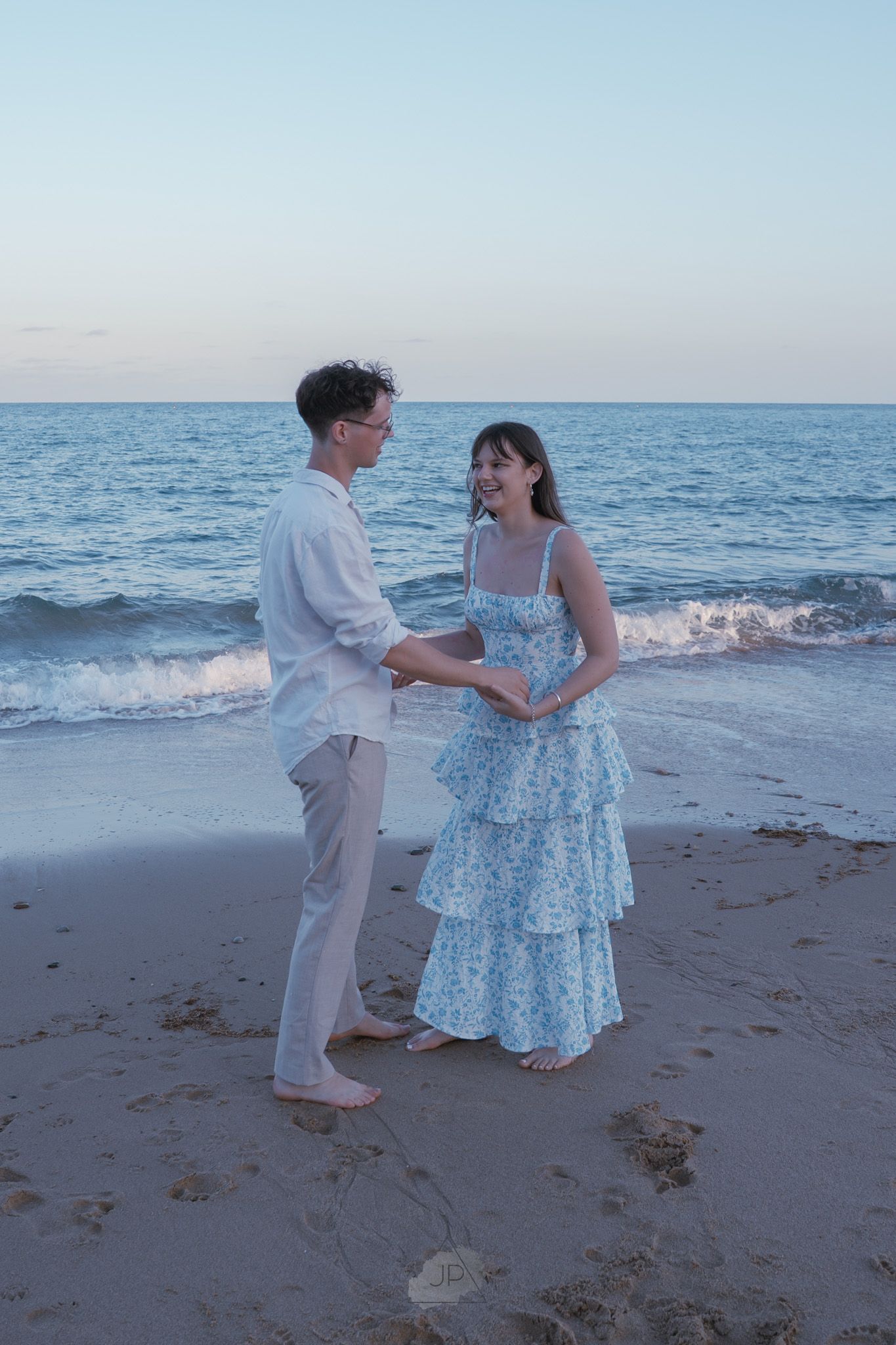A couple dances together on a sandy beach at sunset with gentle ocean waves in the background.