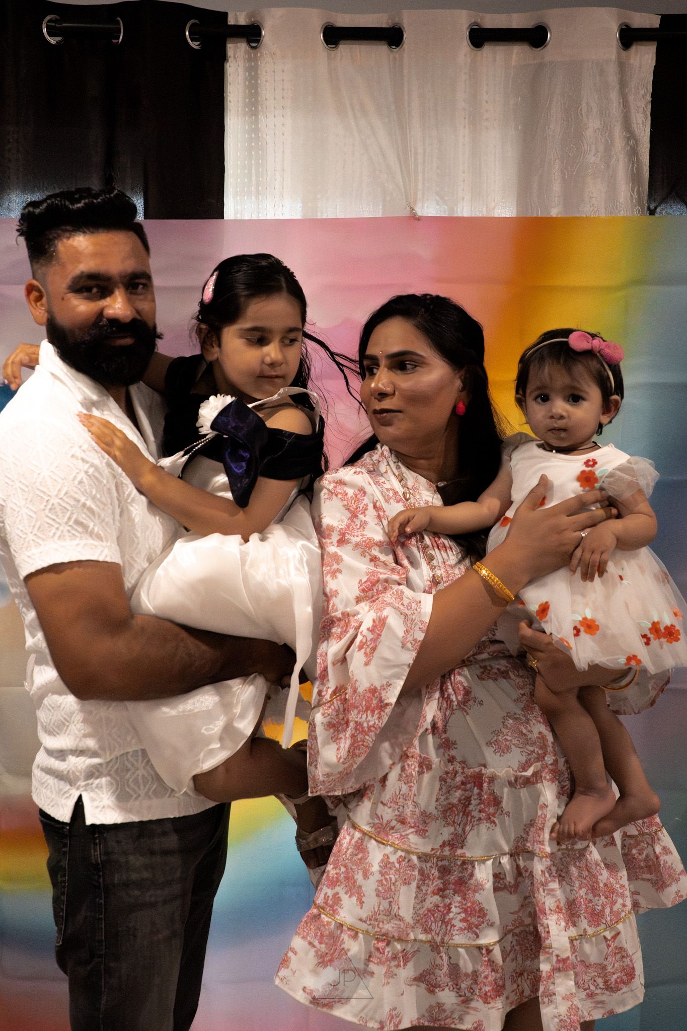 A family poses together in front of a rainbow backdrop wearing coordinated white and floral outfits.