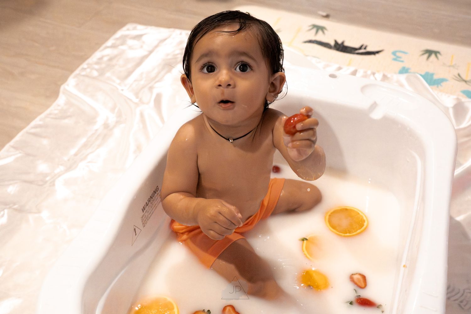 A small child sits in a bathtub playing with orange bath toys while looking upward with curiosity.