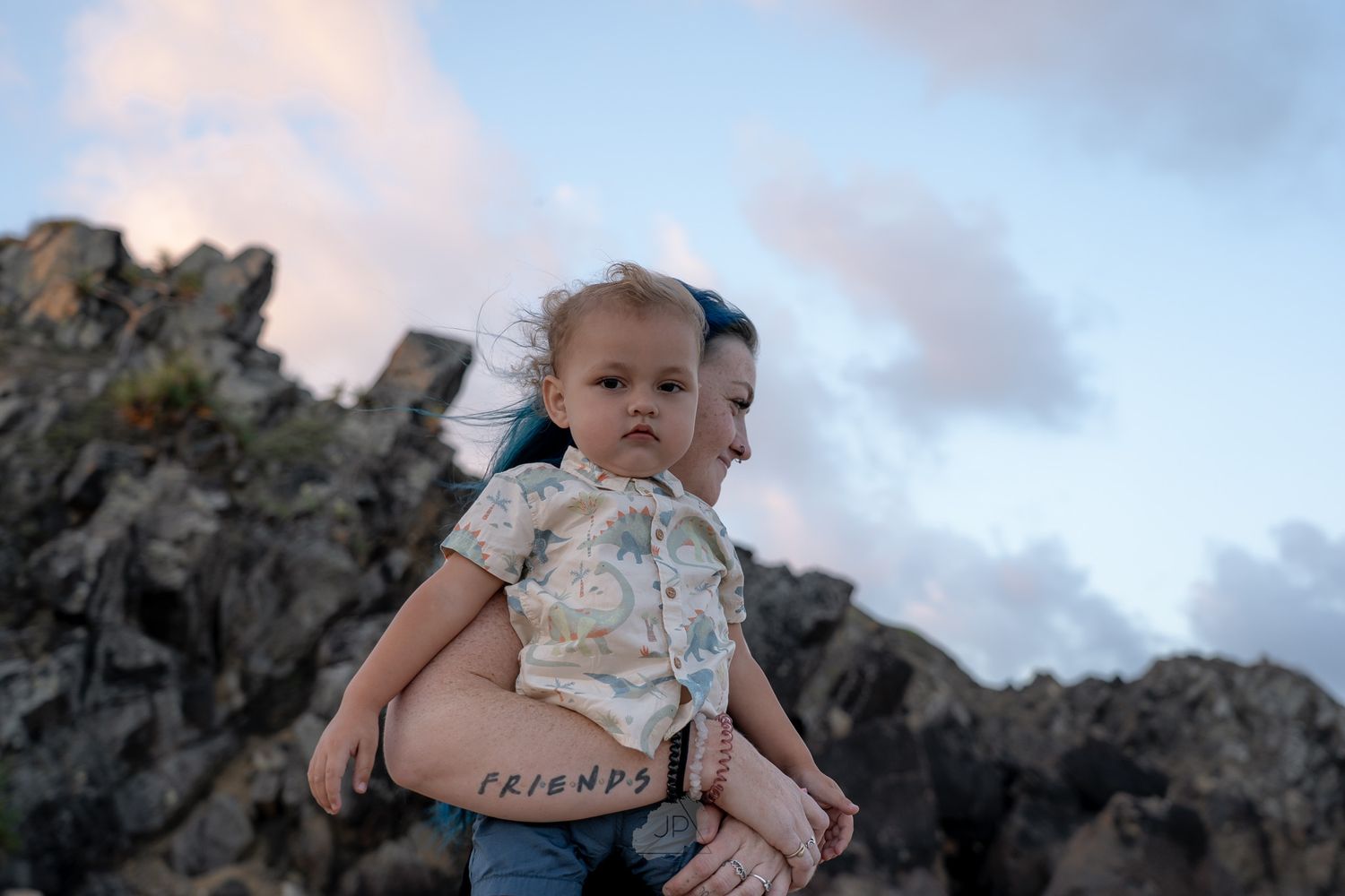 A person in a floral dress holds a small baby while standing against a cloudy sky backdrop.