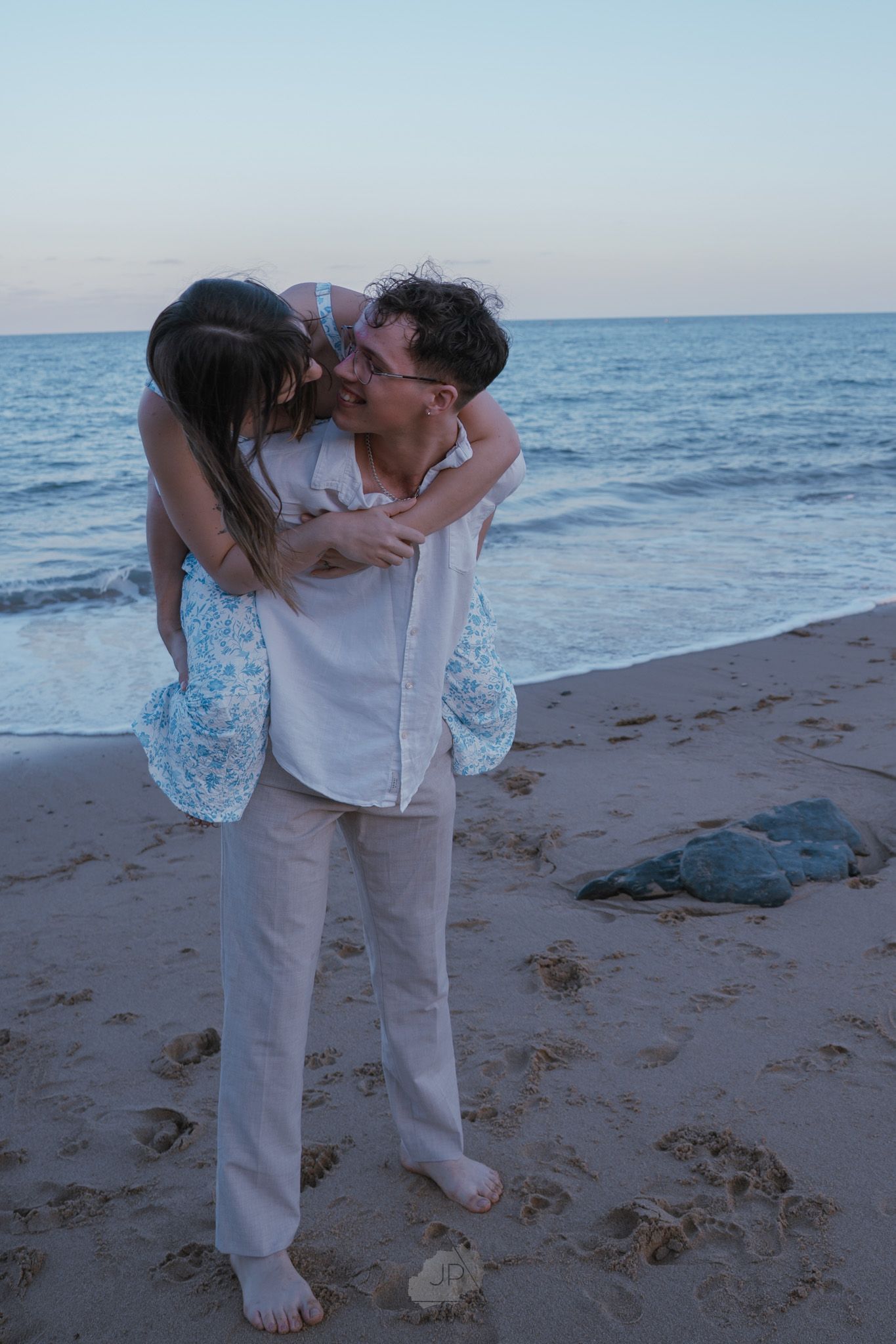 A romantic couple shares a tender kiss on a sandy beach at sunset with ocean waves in the background.