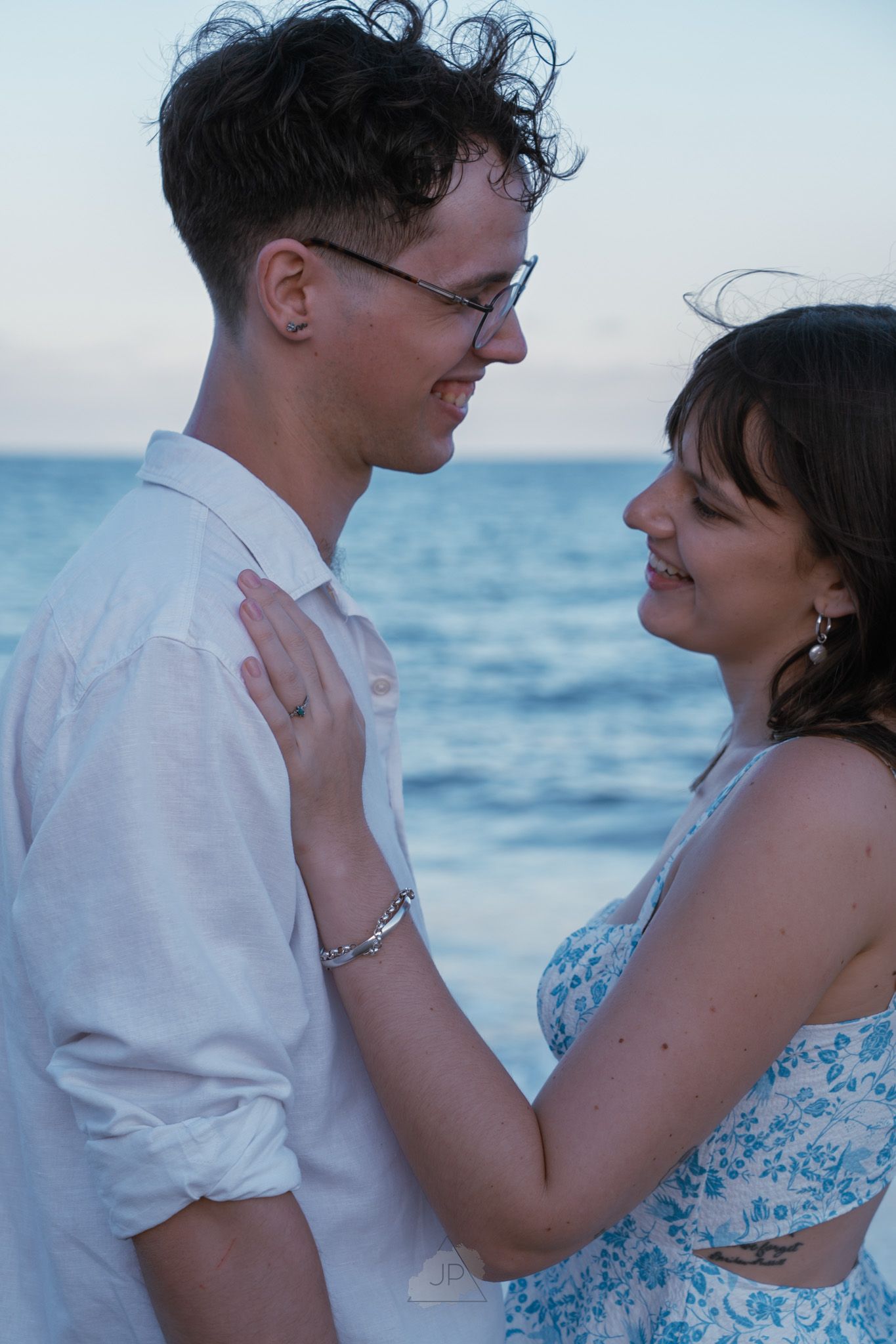 A romantic beachside moment at sunset with a couple embracing in white and blue floral attire against the ocean backdrop.