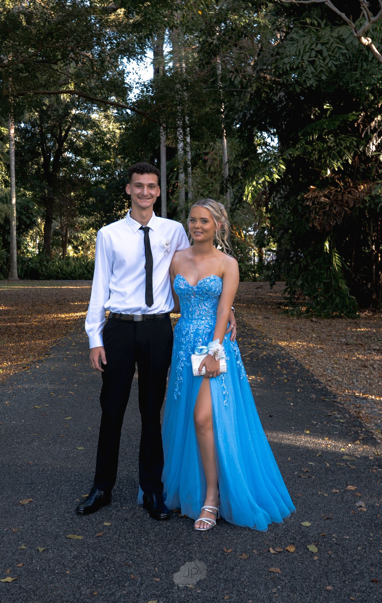 A couple dressed in formal attire stands on a tree-lined path, with the woman wearing a flowing blue gown.