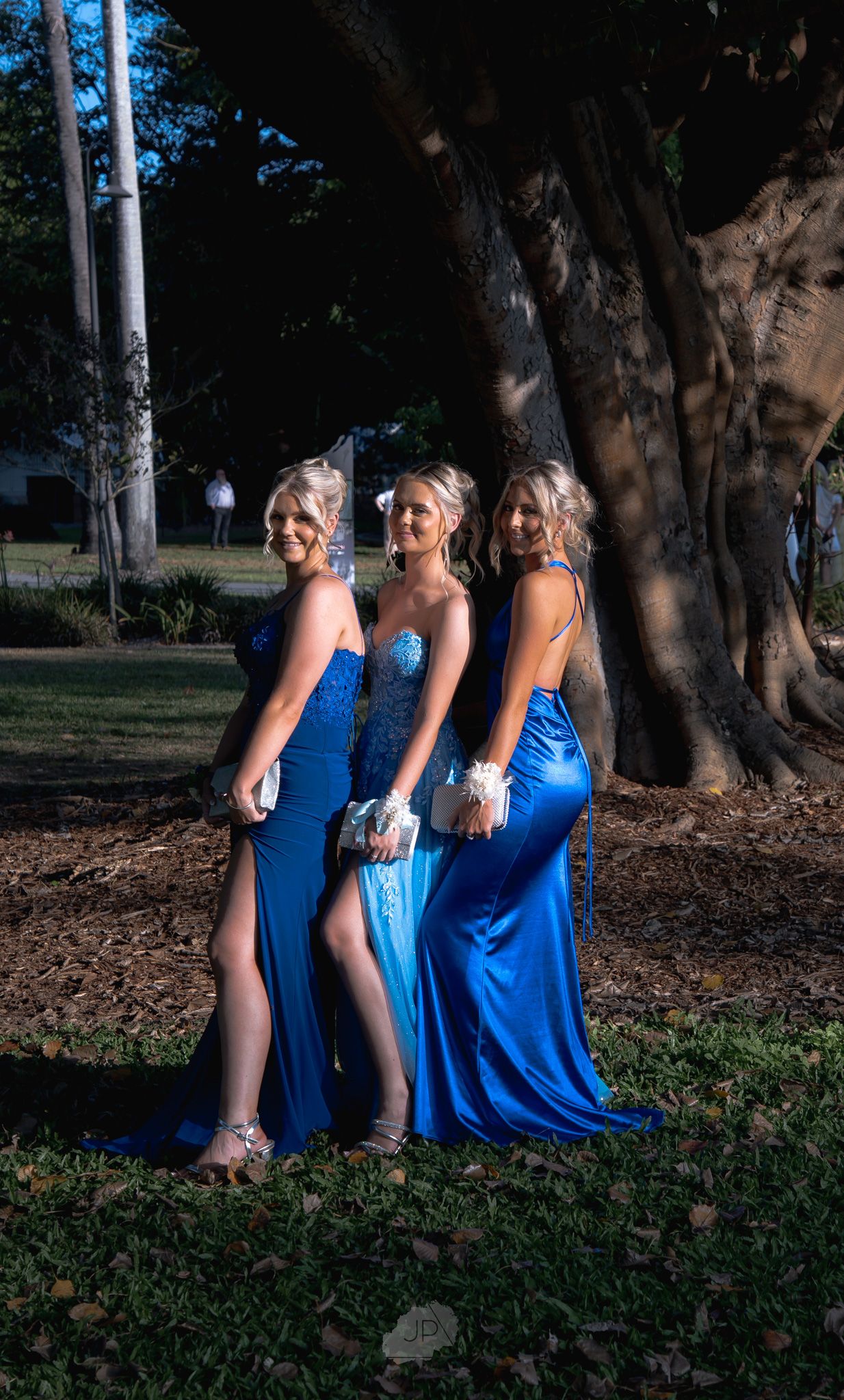 A group in matching blue formal dresses poses playfully in a park setting with trees in the background.