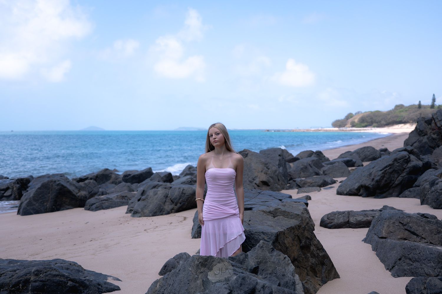 Someone in a pink dress stands among black volcanic rocks on a Hawaiian beach with a lighthouse visible in the distance.