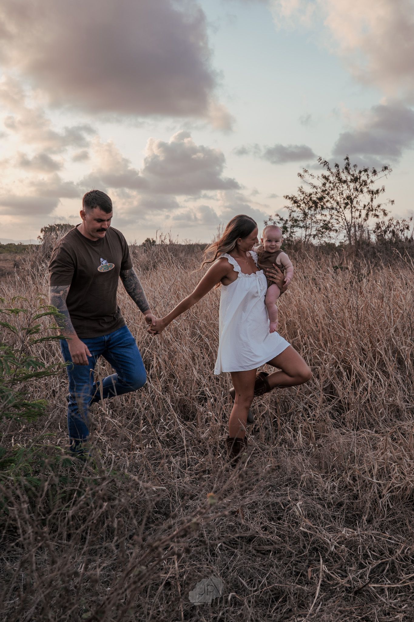 Two people dance and embrace while surrounded by tall dried grass at sunset.