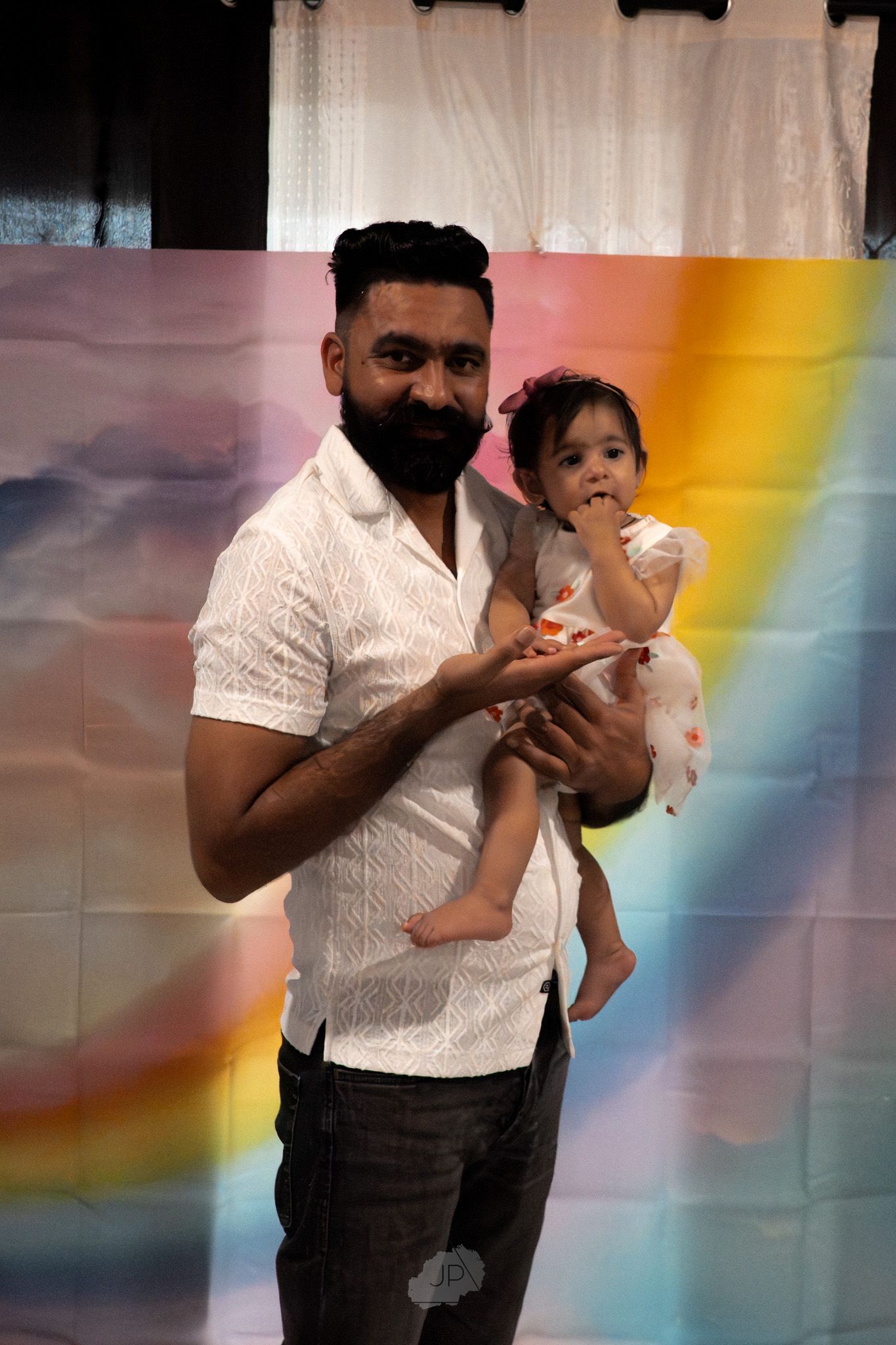 A person in a white shirt holds a small child while standing against a colorful rainbow background.