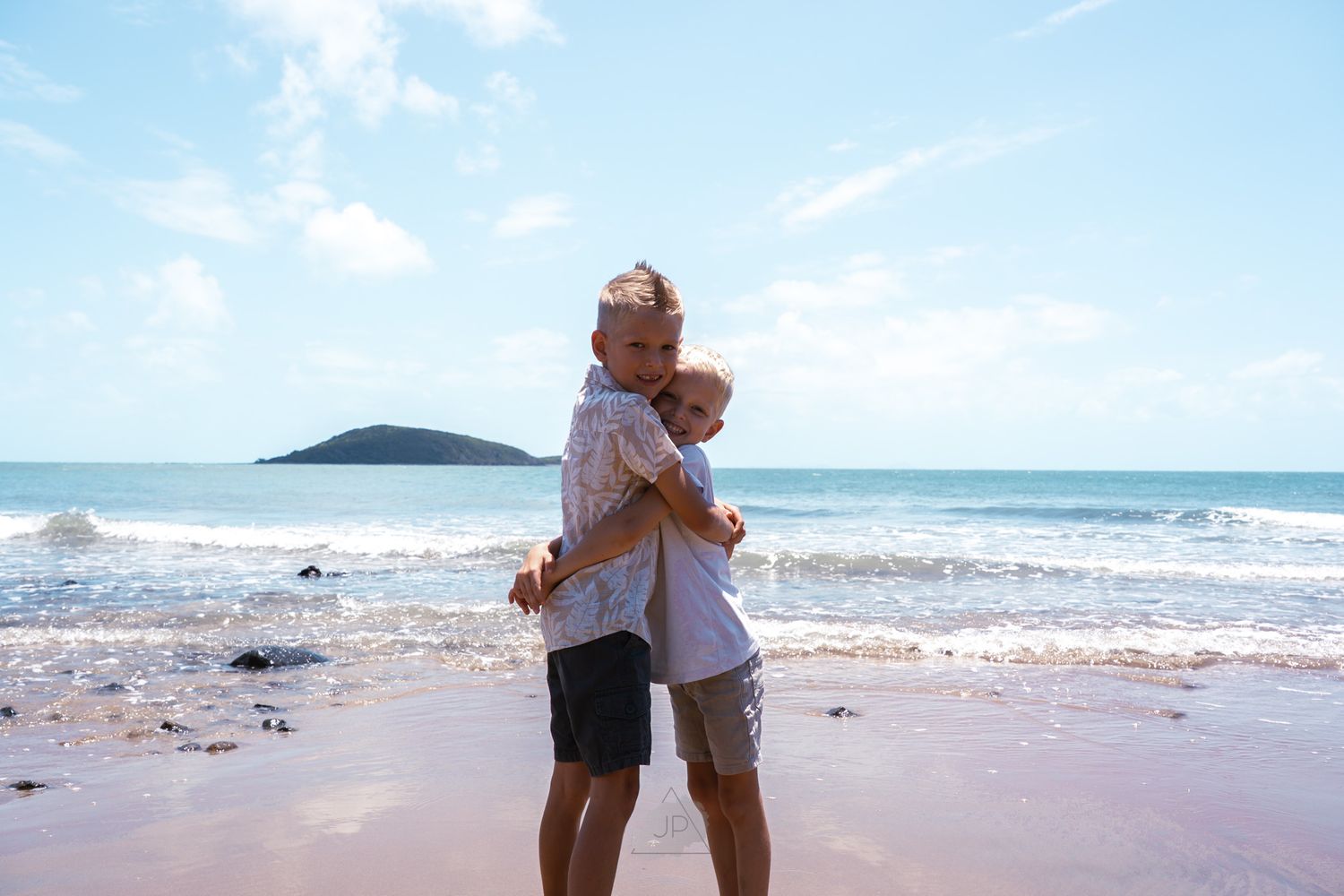 A couple shares a tender moment while standing on a tropical beach with ocean waves and distant island views.