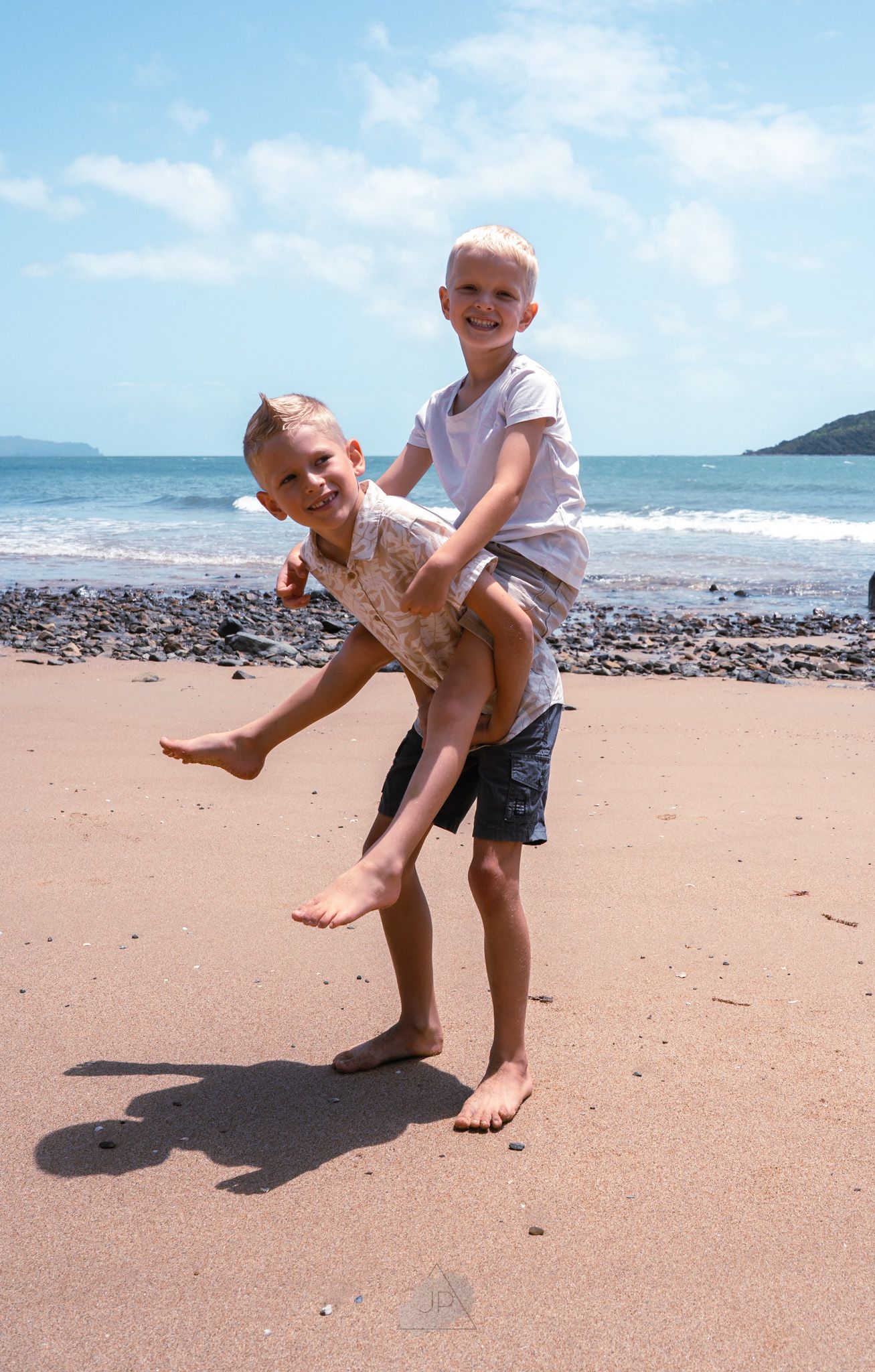 Two children play together on a sandy beach with ocean waves and mountains visible in the background.