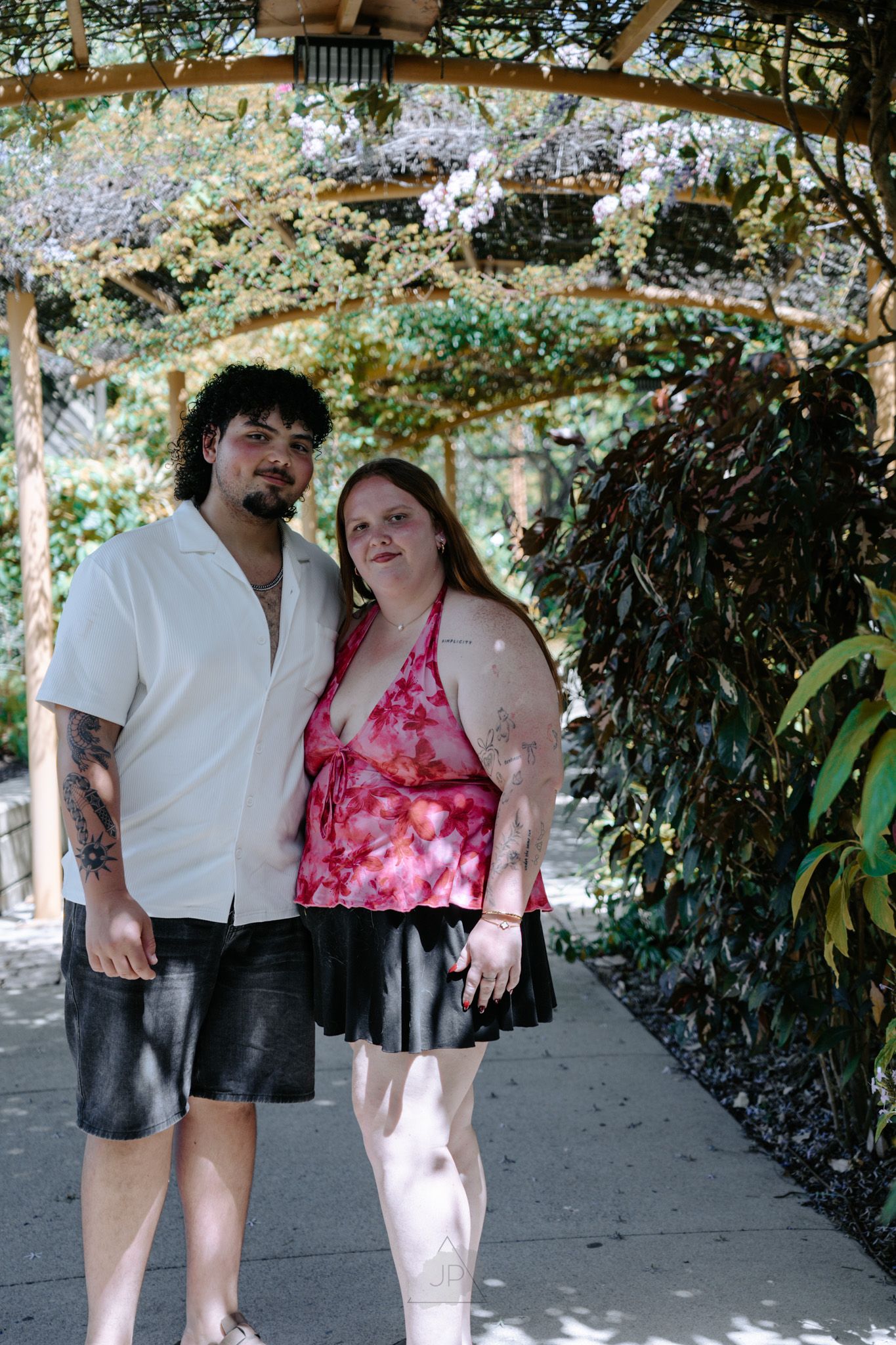 A couple stands together under a shaded garden pathway in summer attire.