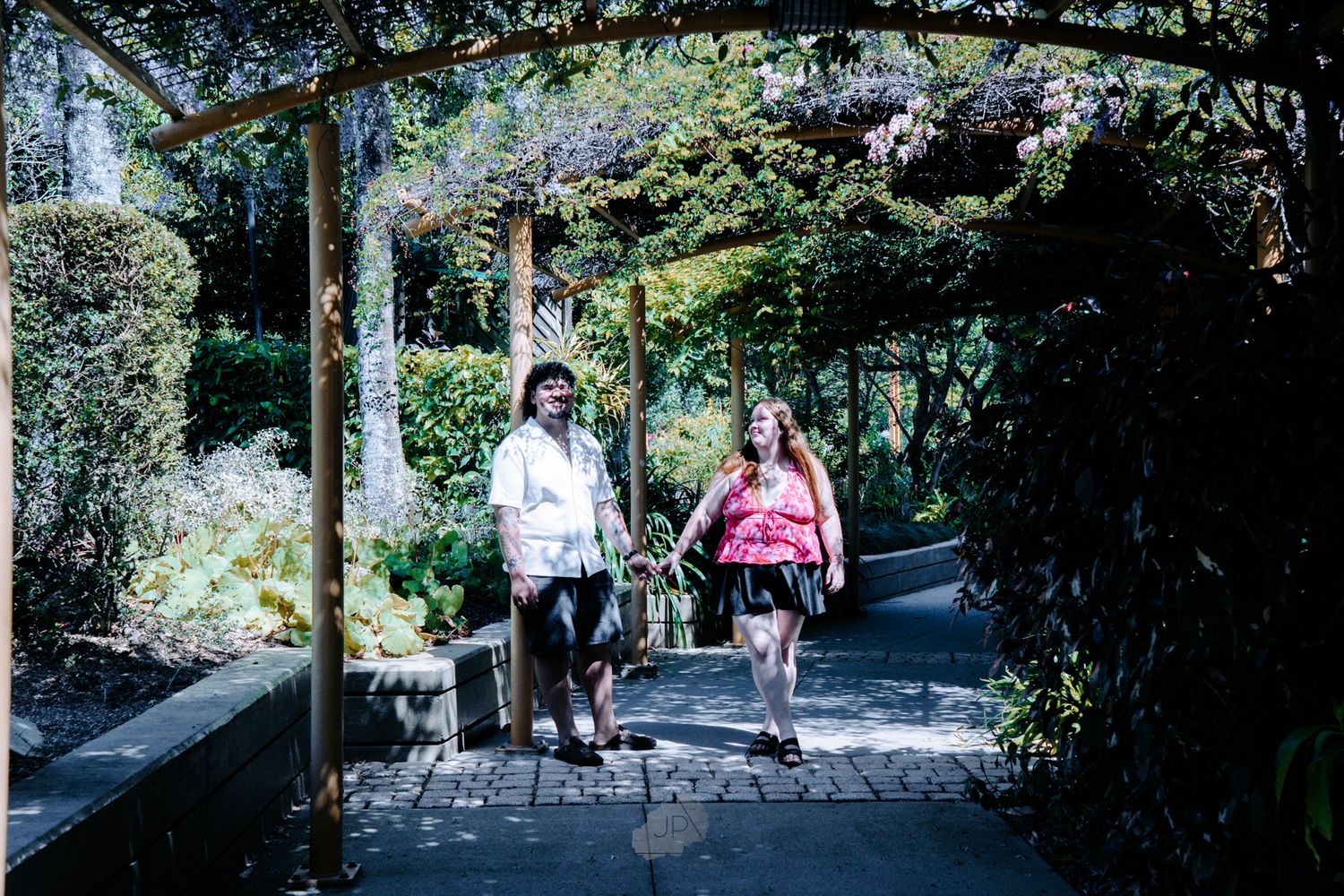 Two people walking dogs on a shaded garden path surrounded by lush greenery and trees on a sunny day.