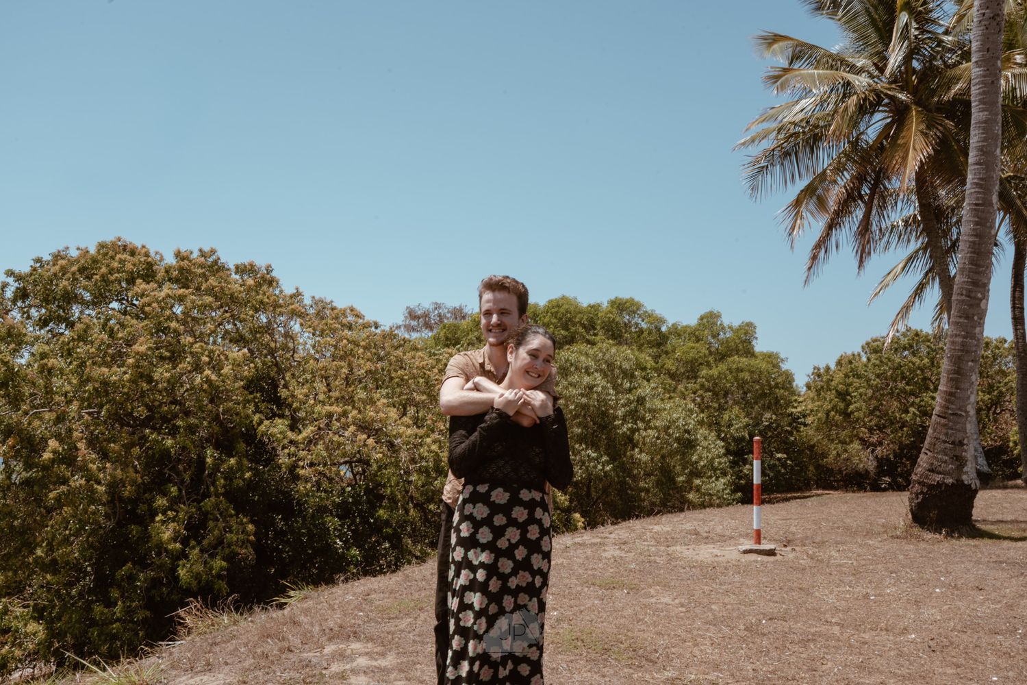 A person in a polka dot dress stands on a grassy hill with palm trees and blue sky in the background.