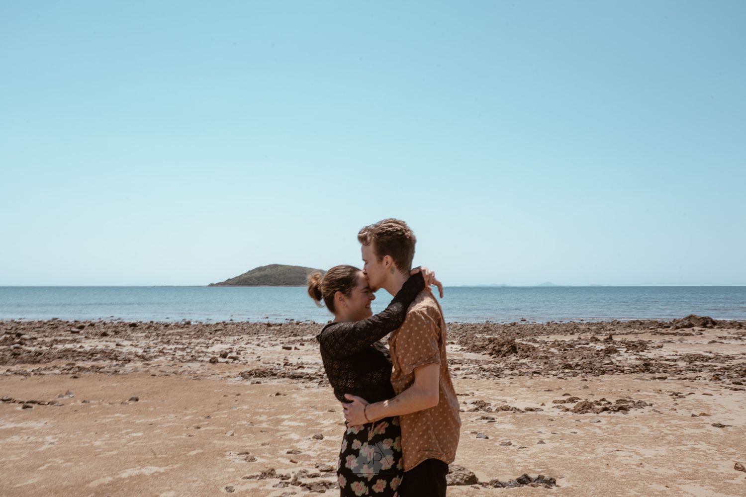 A couple embraces on a sandy beach with a small island visible on the horizon under a bright blue sky.