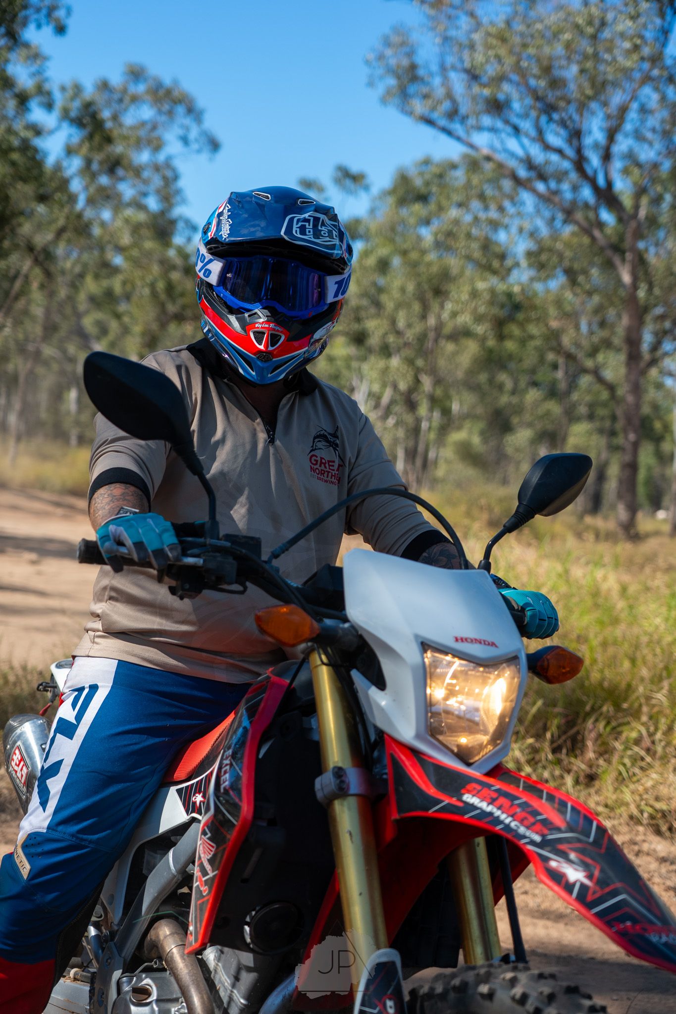 A dirt bike rider in blue gear navigating a trail through bushland on a red motorcycle.