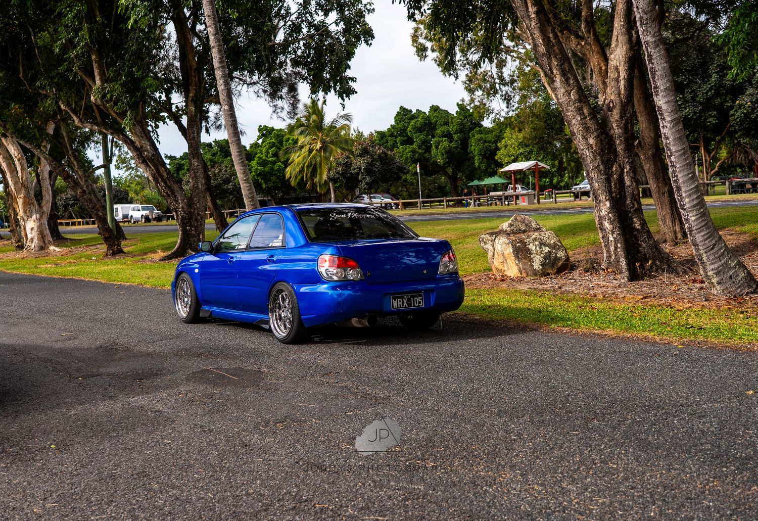 A bright blue Subaru WRX STI sports car parked on a tree-lined path with dappled sunlight filtering through.