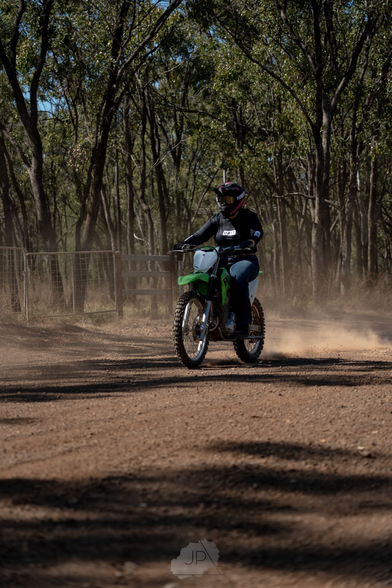 A motorcycle rider navigates through a dusty forest trail, leaving a cloud of dirt in their wake.