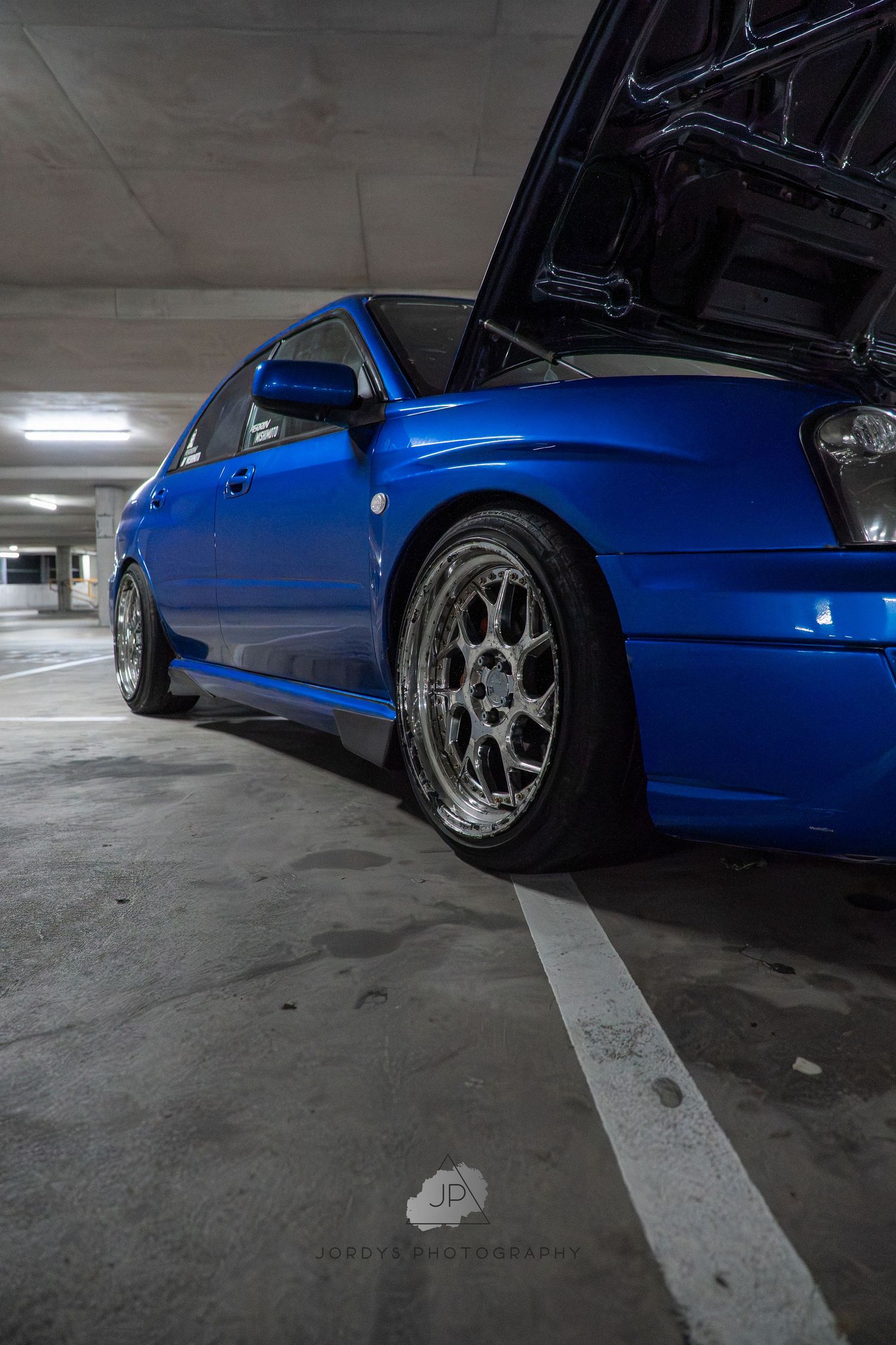 Low angle shots of a bright blue sports car with custom silver wheels parked in a concrete parking garage.