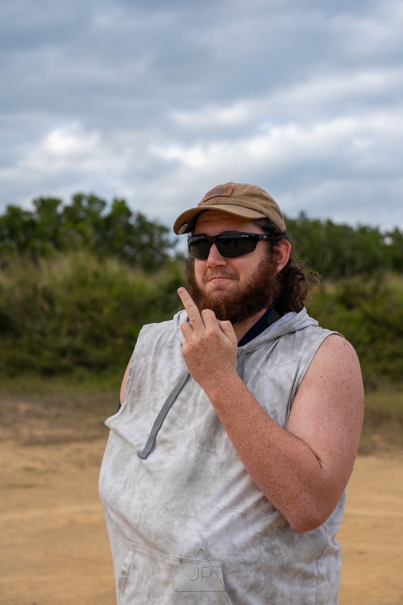 Person wearing sunglasses and sleeveless shirt stands outdoors under cloudy sky in sandy area.