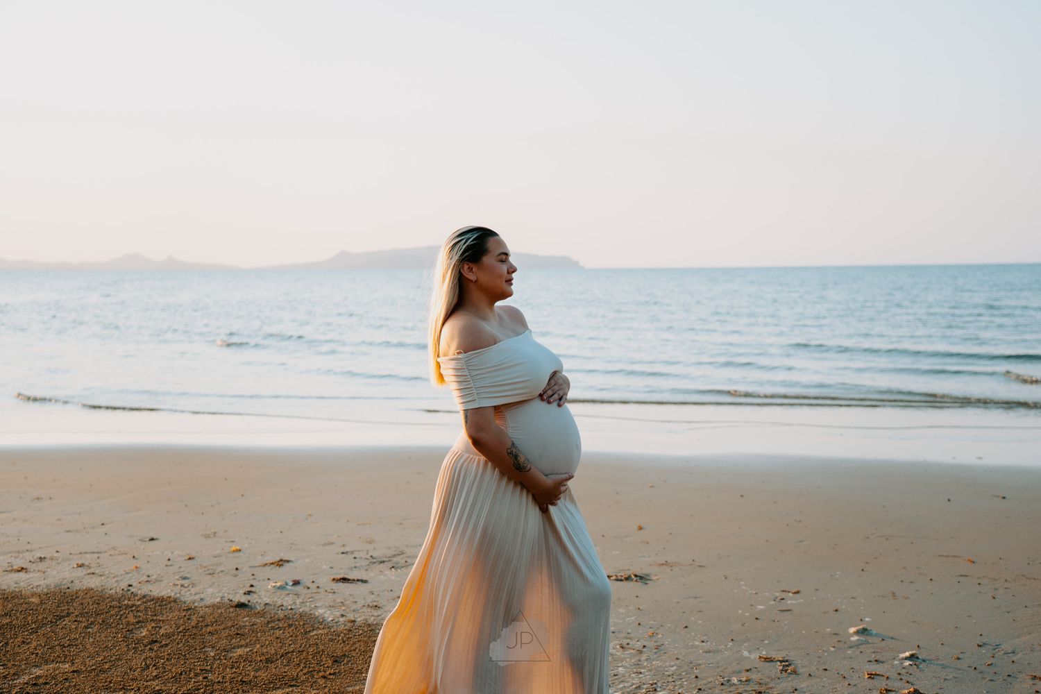A pregnant woman in a white dress walks along a serene beach at sunset with ocean waves in the background.