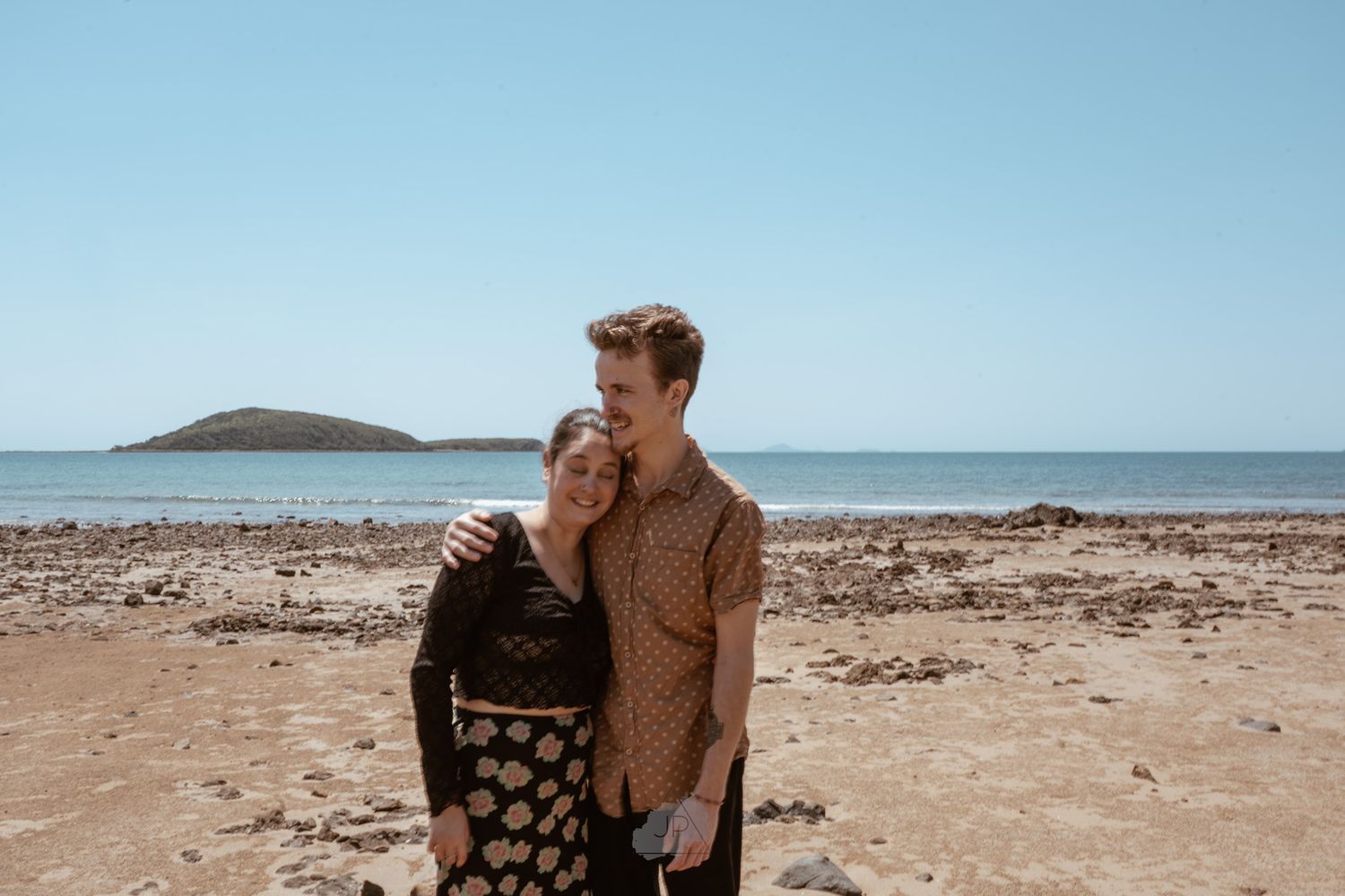 A couple embraces while walking along a scenic desert coastline with blue ocean waters and clear skies in the background.