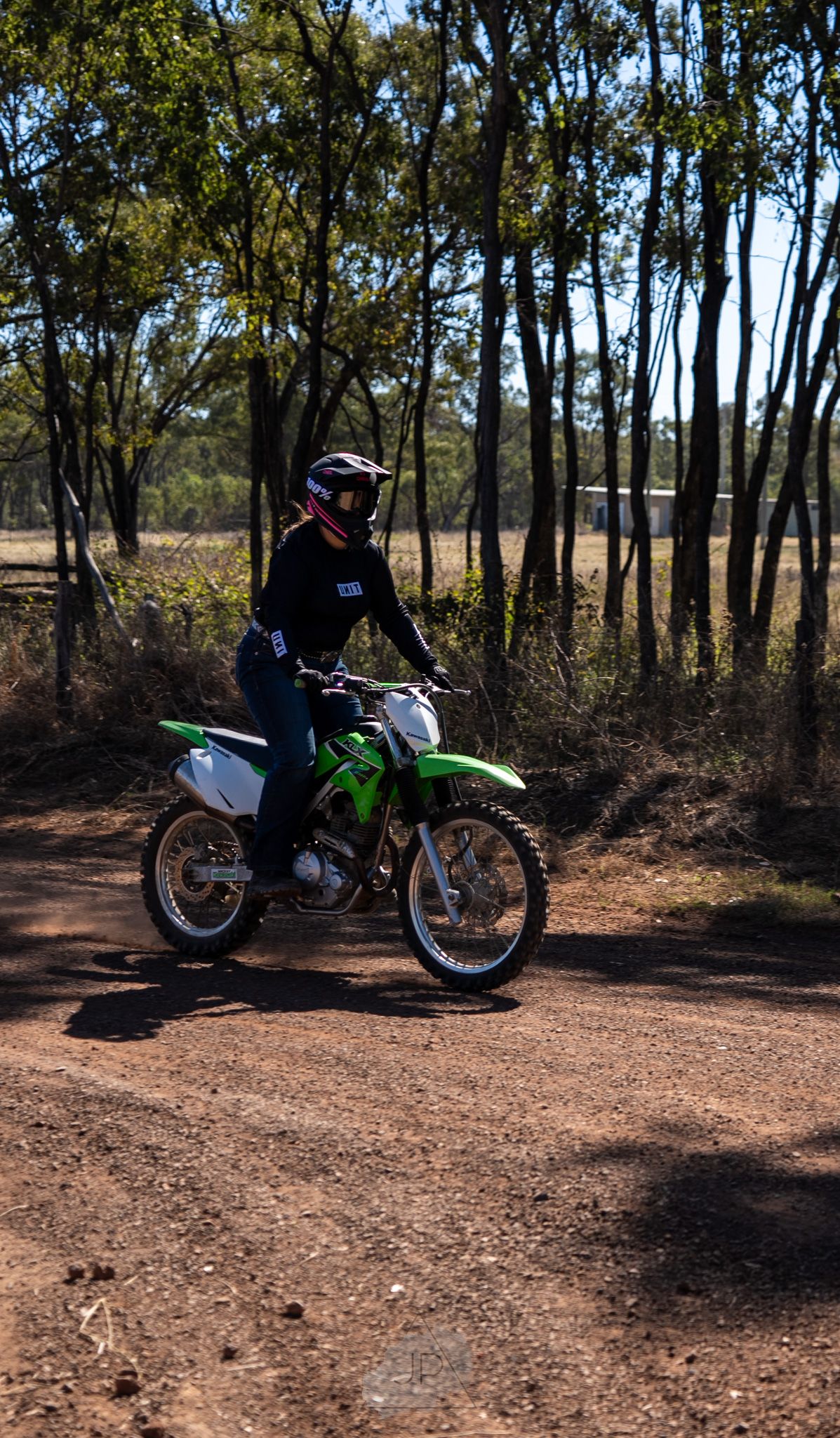 A green dirt bike and rider navigating through a wooded area on a dirt trail with trees in the background.