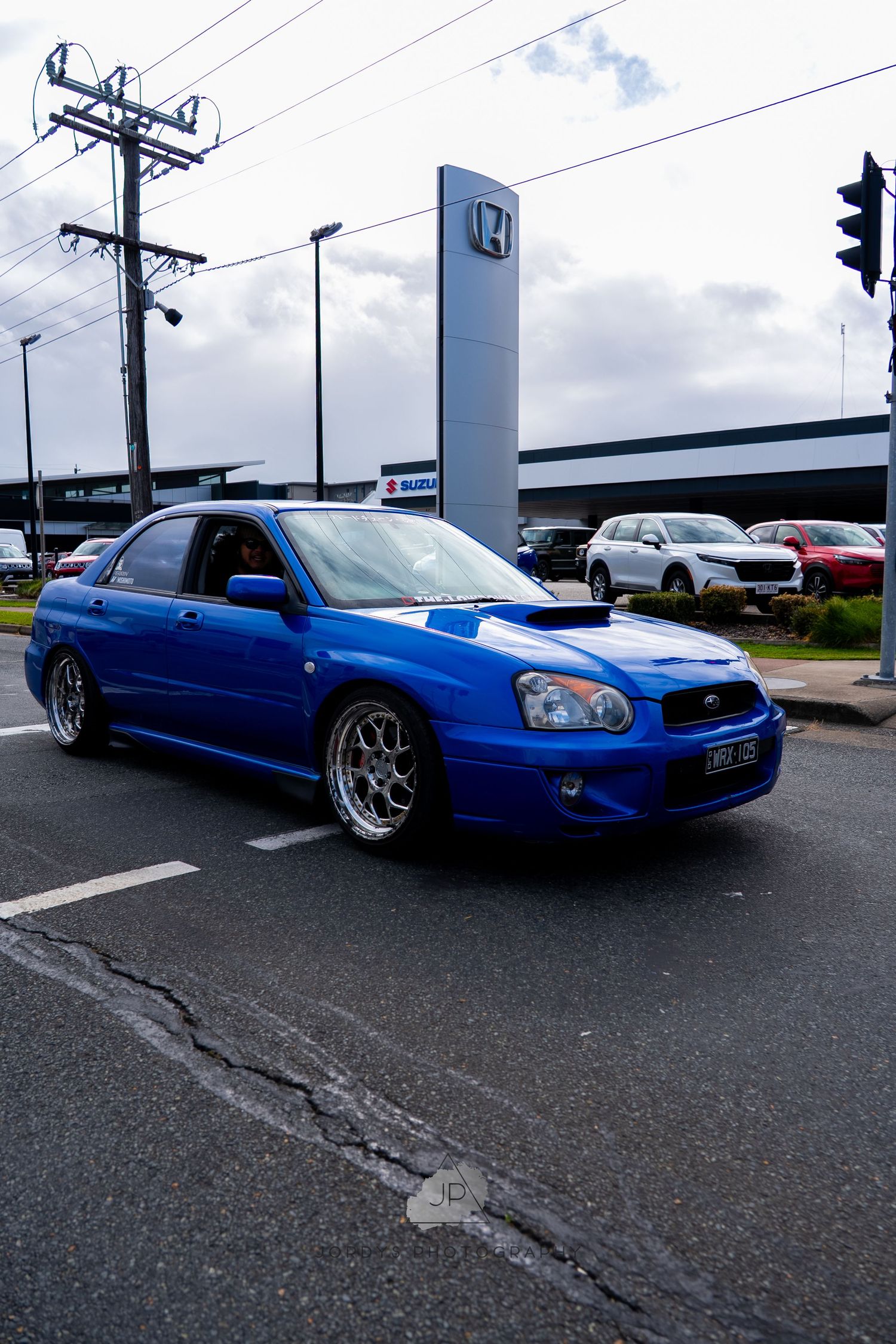 A modified blue Subaru WRX with lowered suspension and aftermarket wheels parked in a dealership lot.