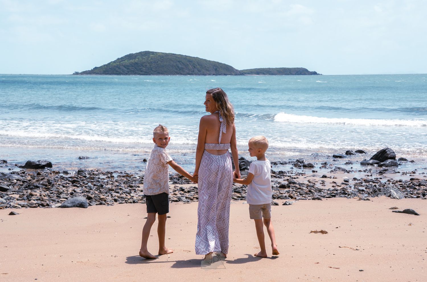 Three people walking along a scenic beach shoreline with rocky outcrops and an island visible in the distance.