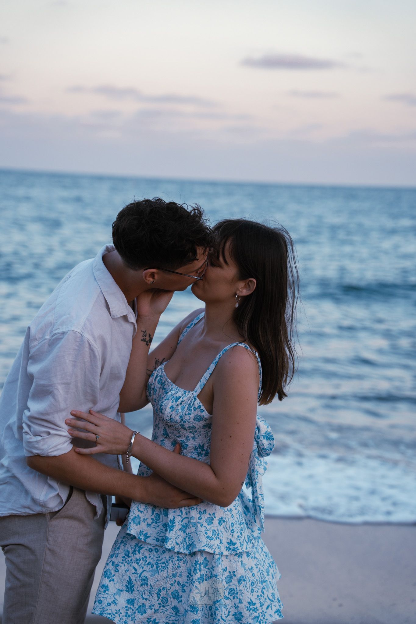 A romantic couple shares tender kisses on a serene beach at sunset with ocean waves in the background.