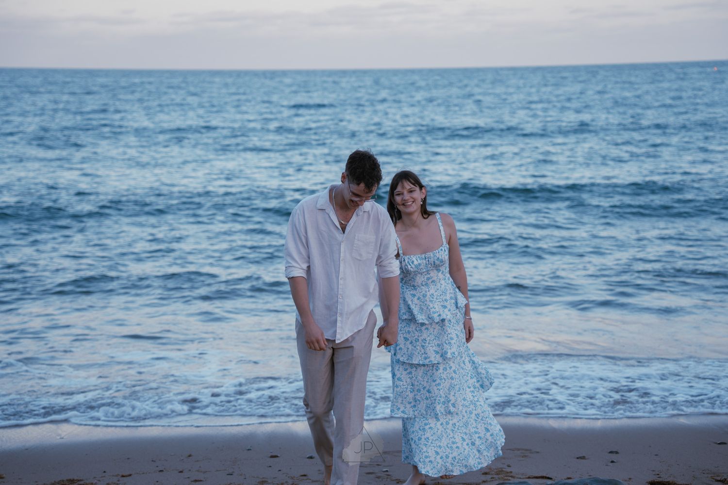 Couple walking along a scenic beach at sunset with ocean waves in the background.