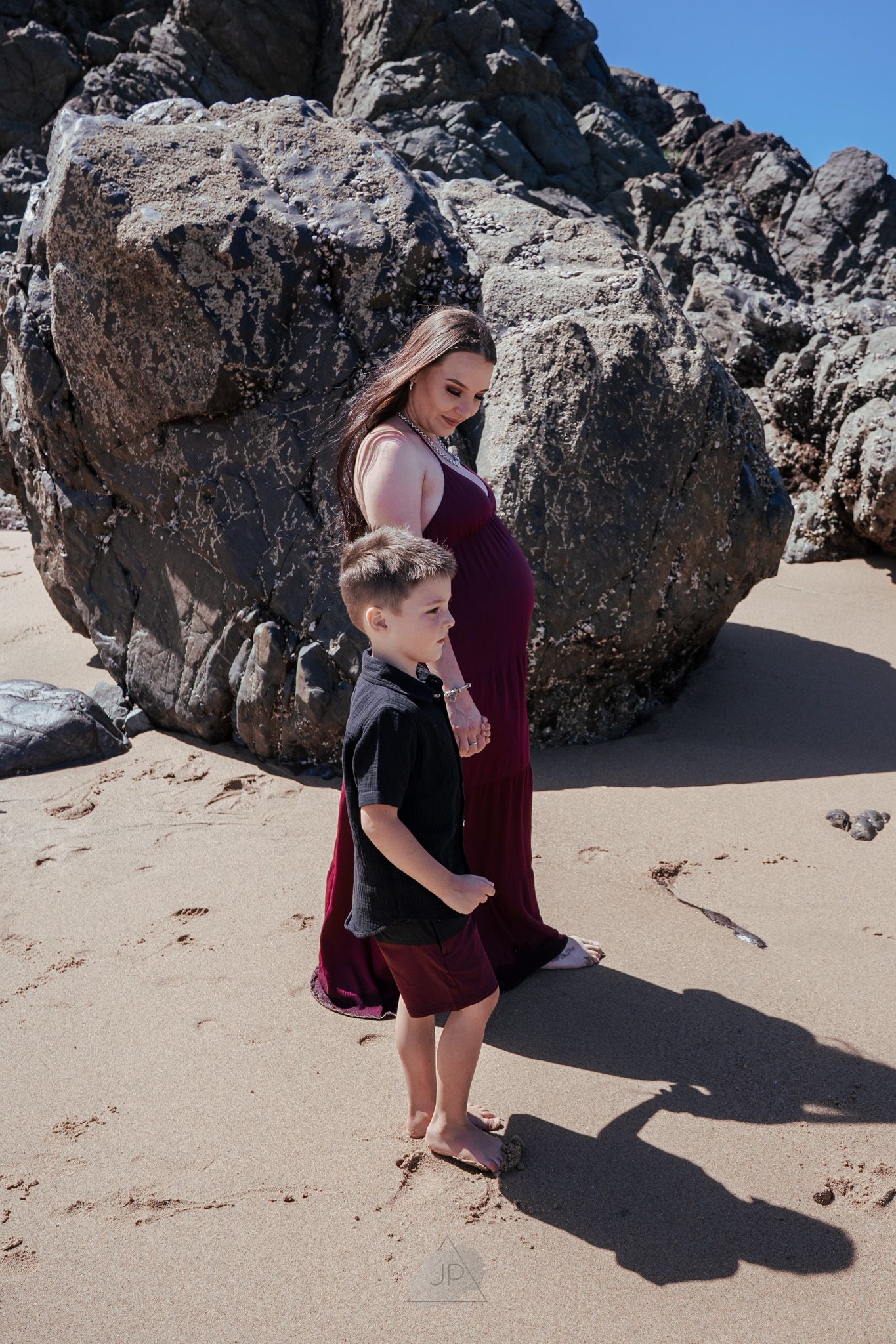 A person in burgundy clothing explores rocky formations on a sandy beach during a sunny day.
