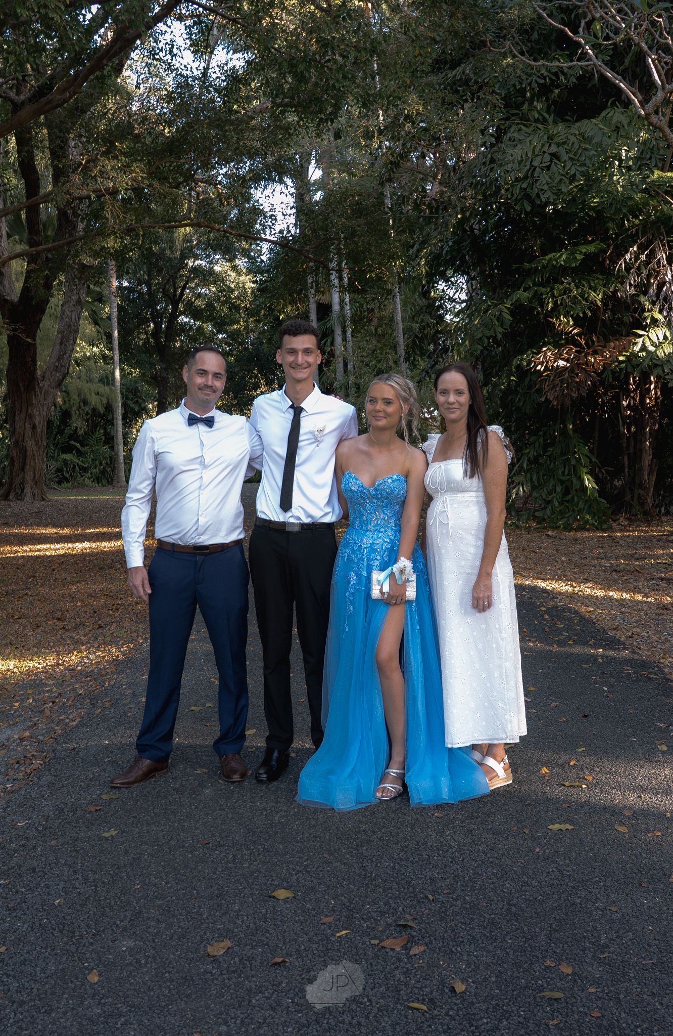 A group in formal attire poses outdoors on a path surrounded by trees during an autumn day.
