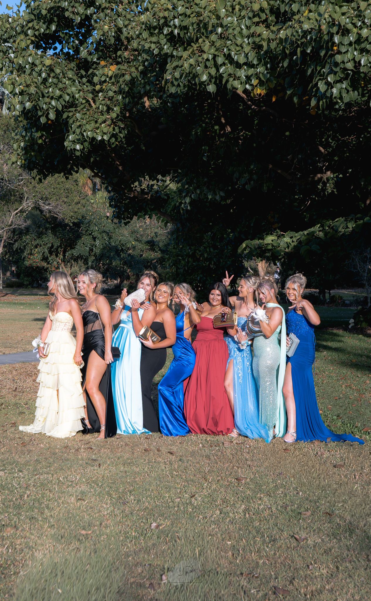 A group wearing formal dresses in shades of blue and burgundy poses together outdoors on a grassy lawn.