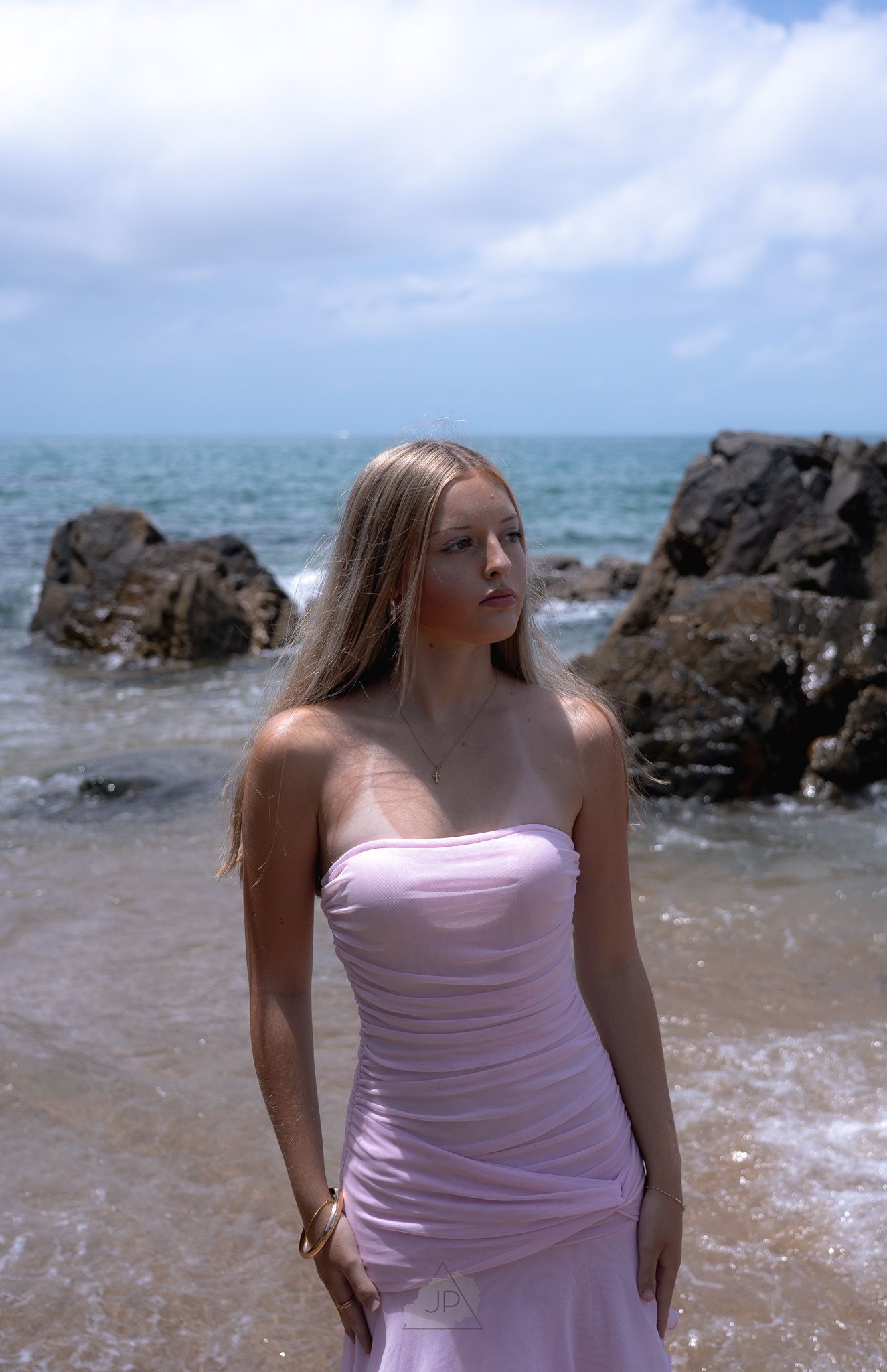 A person in a pink dress poses on a rocky beach with ocean waves and cloudy sky in the background.