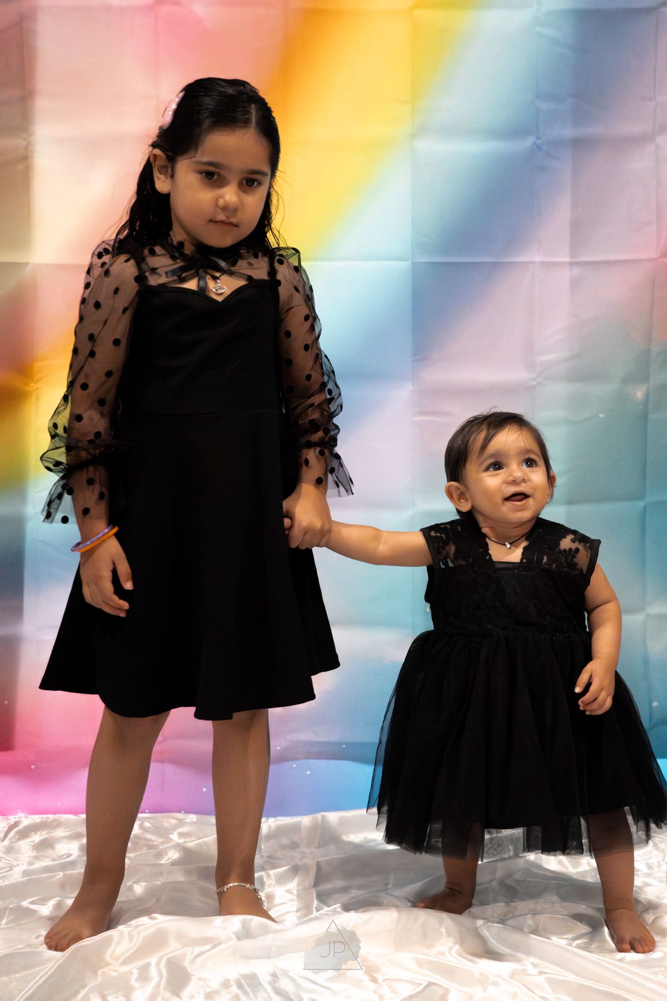 Two figures in matching black lace dresses pose together against a colorful rainbow backdrop.