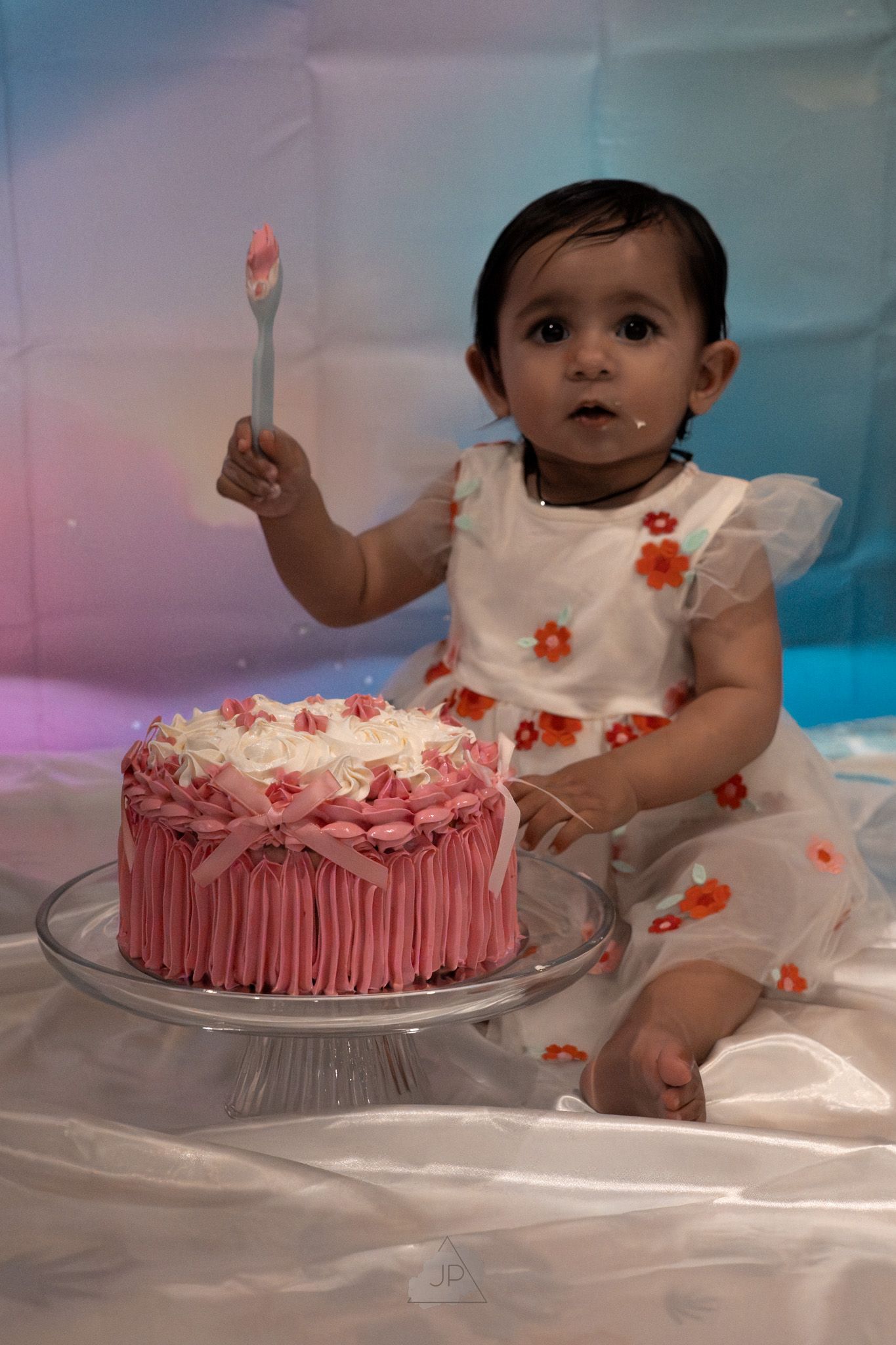 A small child in a white floral dress reaches toward a pink frosted cake on a glass stand.