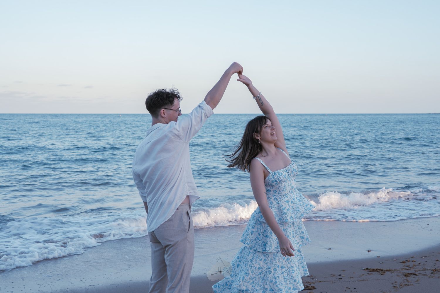 Couple twirling and dancing playfully along the shoreline as waves roll in during golden hour.