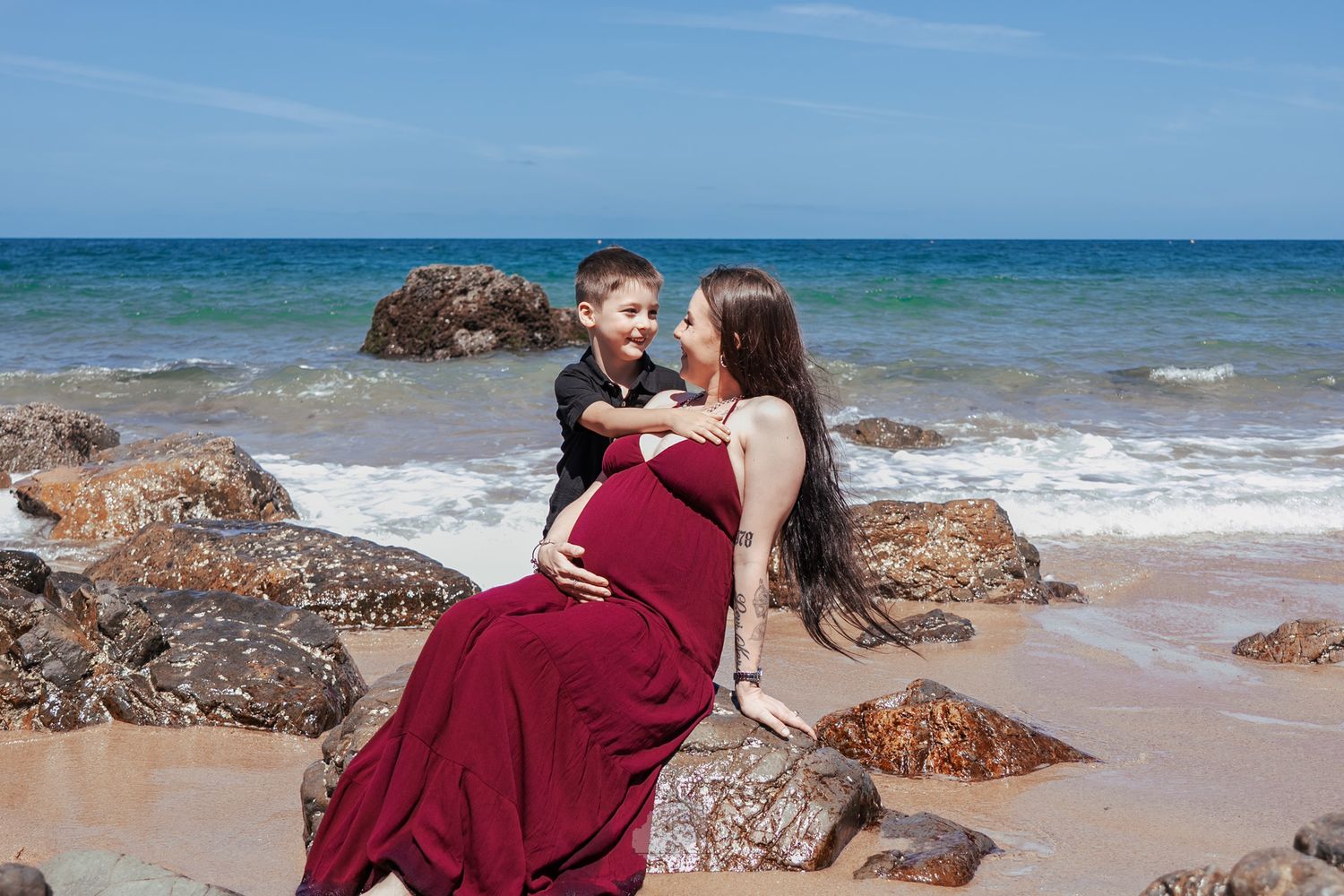 Couple in burgundy attire embracing on a scenic beach with rocky outcrops and turquoise ocean waters.