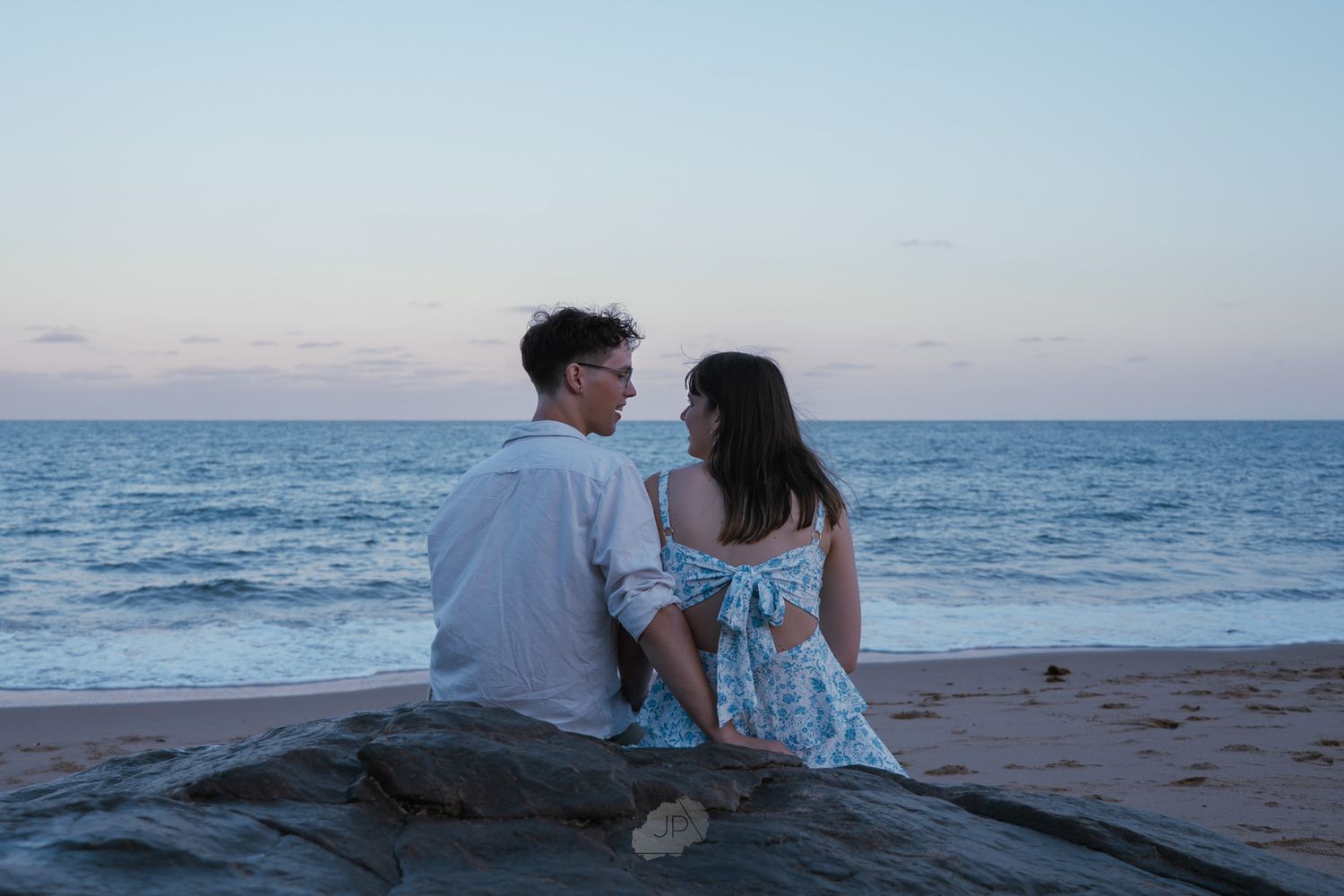 A couple sits together on a beach watching the sunset over the ocean horizon.