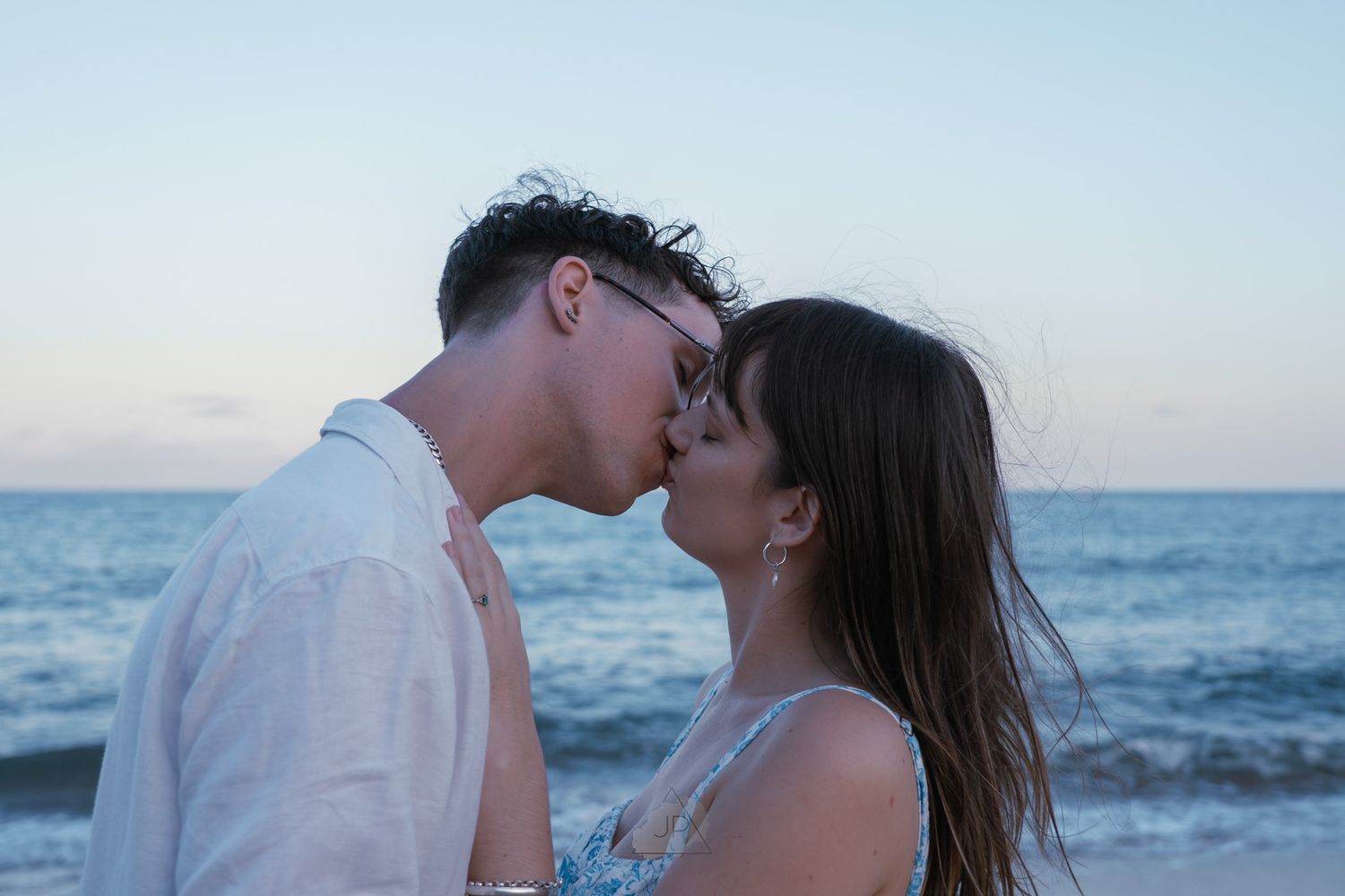 A couple shares a romantic moment at the beach during sunset with waves crashing behind them.
