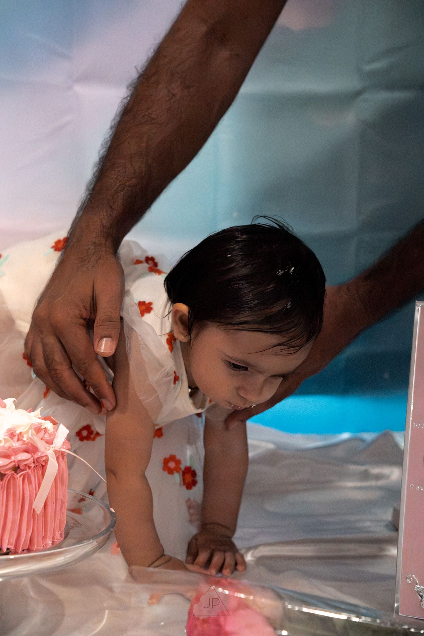 Hands gently guide a small baby through a smash cake photoshoot with pink frosting against a blue backdrop.